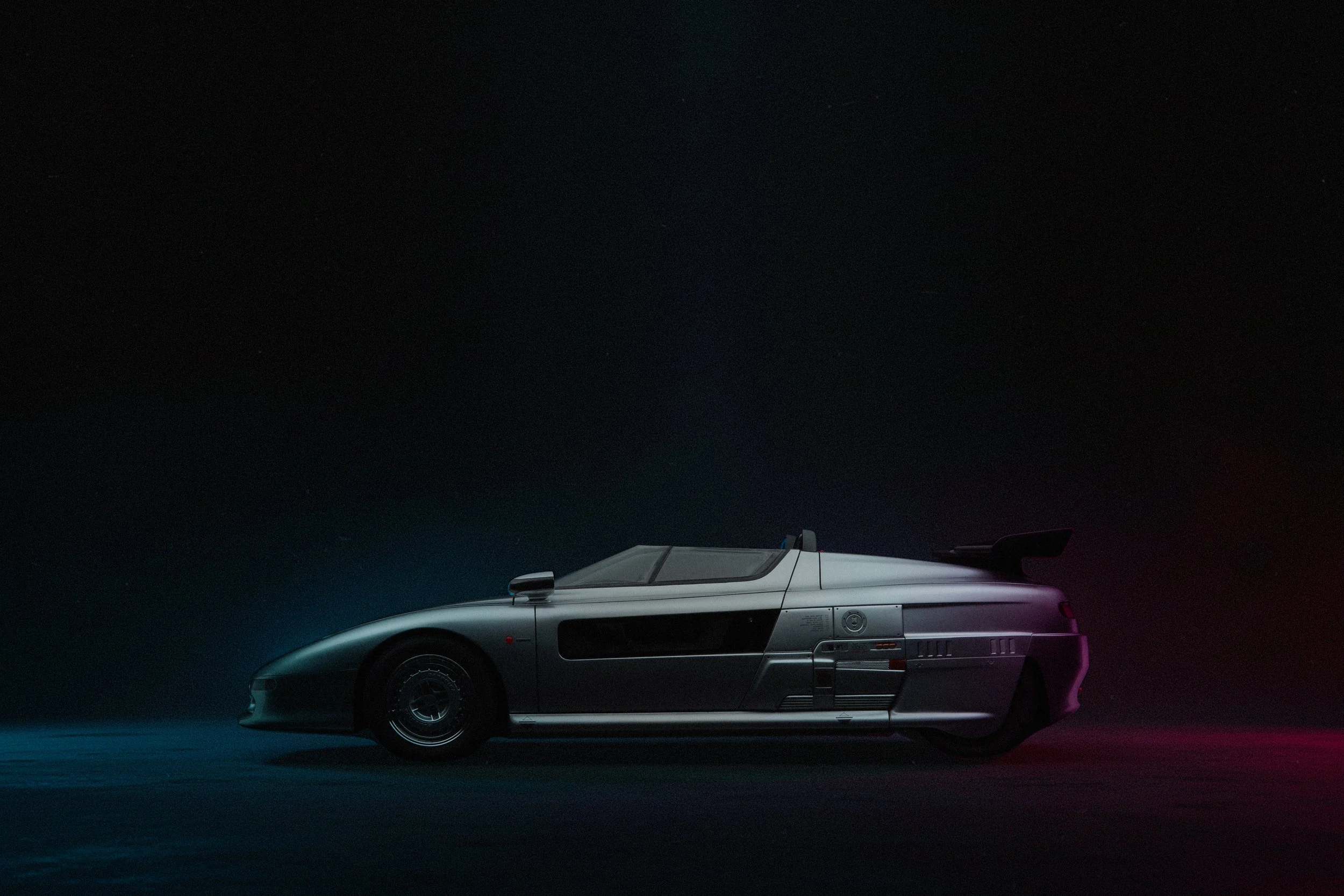 A silver futuristic sports car with a low profile, aerodynamic design, and rear wing, photographed at night with neon lighting.