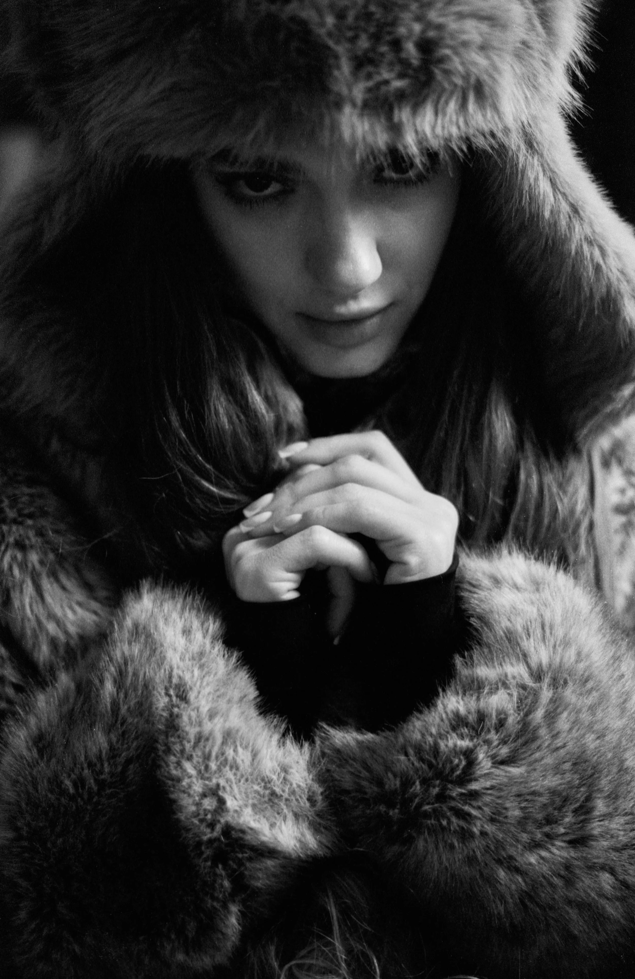 A woman in a fur hood looking into the camera with a serious expression, hands clasped together.