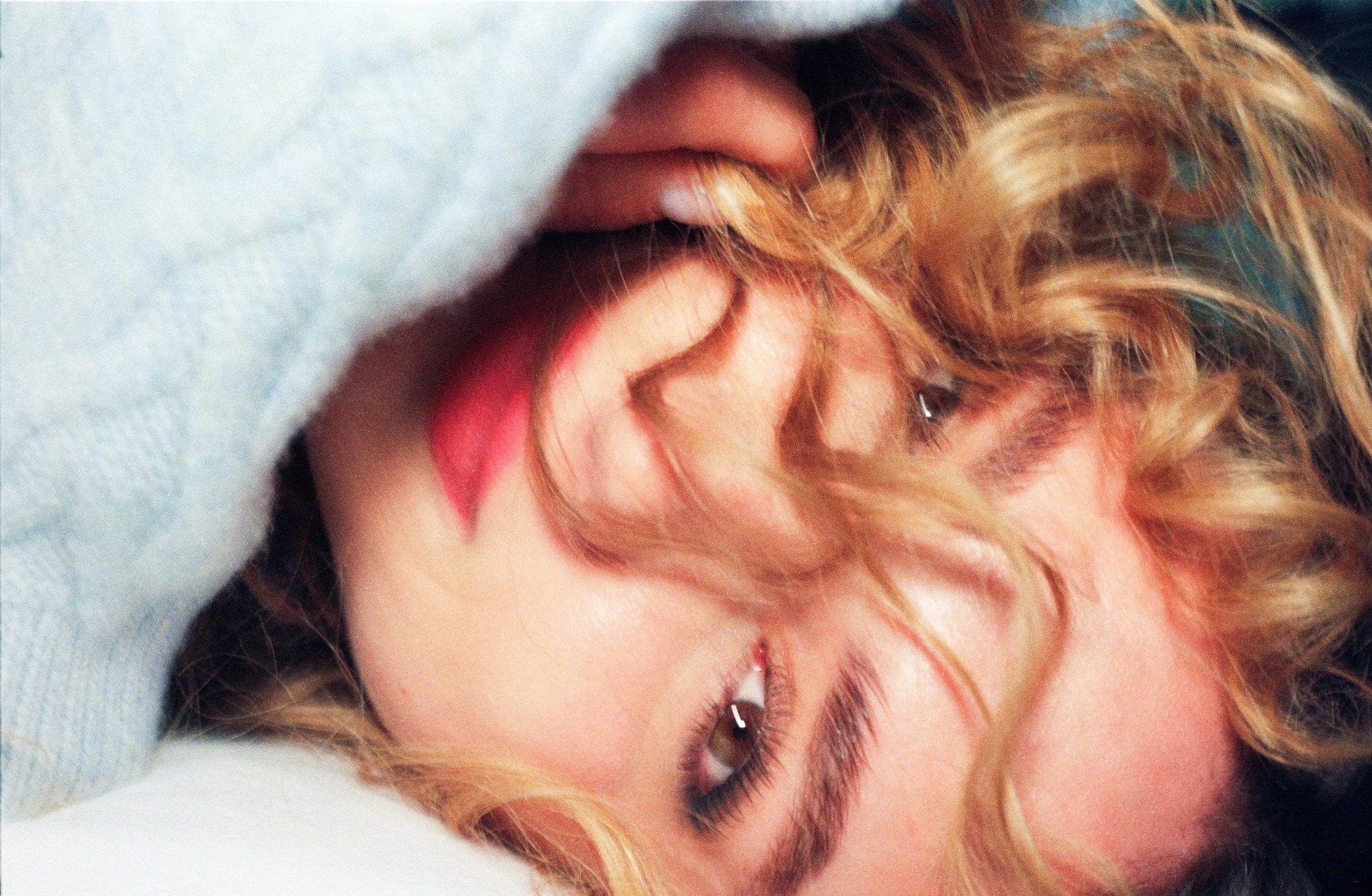 Close-up of a woman with curly blonde hair and makeup, lying down with her eyes closed and a smile.