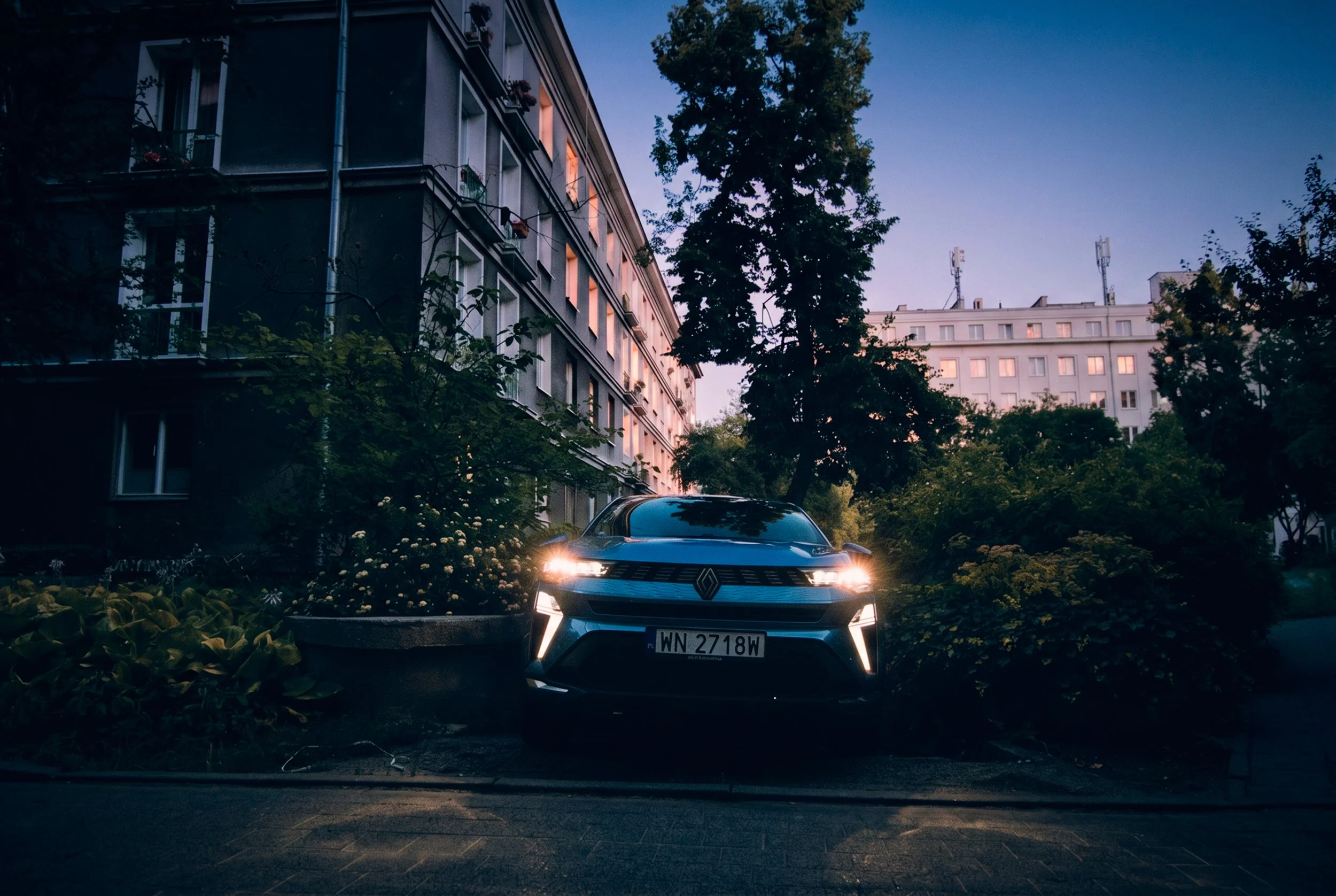A modern electric Renault car with its headlights on, parked in a residential area at dusk, with tall apartment buildings and trees in the background.