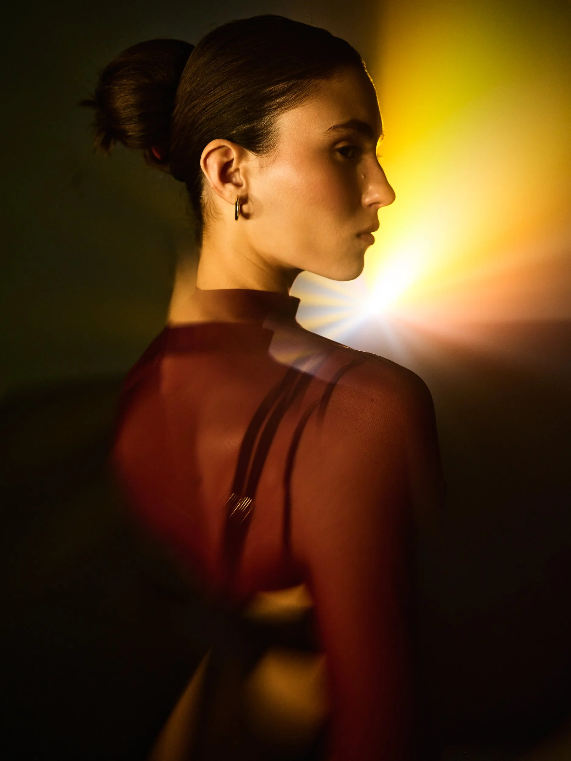 Side profile of a woman with dark hair in a bun, wearing a hoop earring, illuminated by yellow and blue lights.