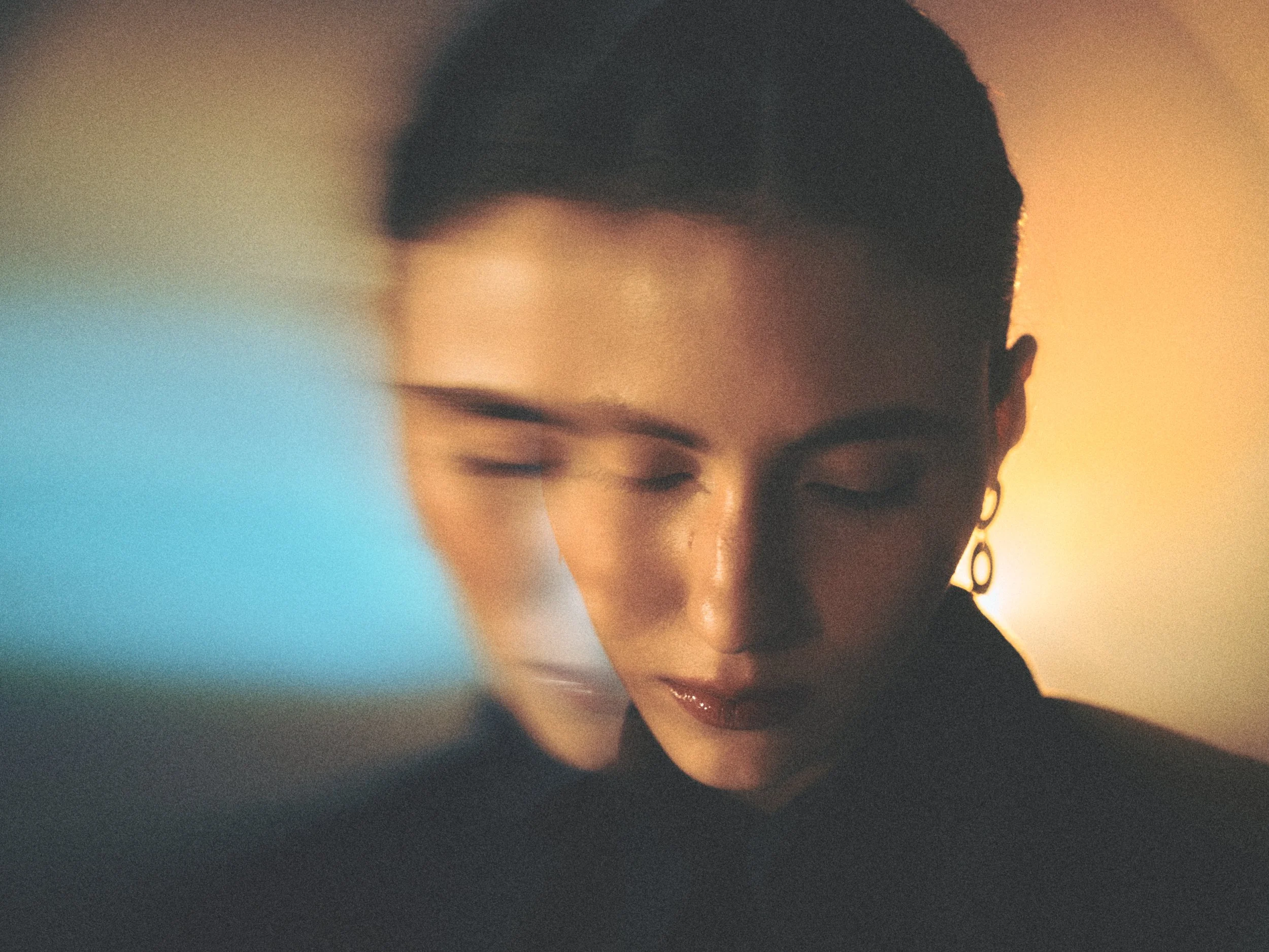 A person with short hair and hoop earrings, looking down with eyes closed, illuminated by warm and cool colored lighting in a dimly lit setting.