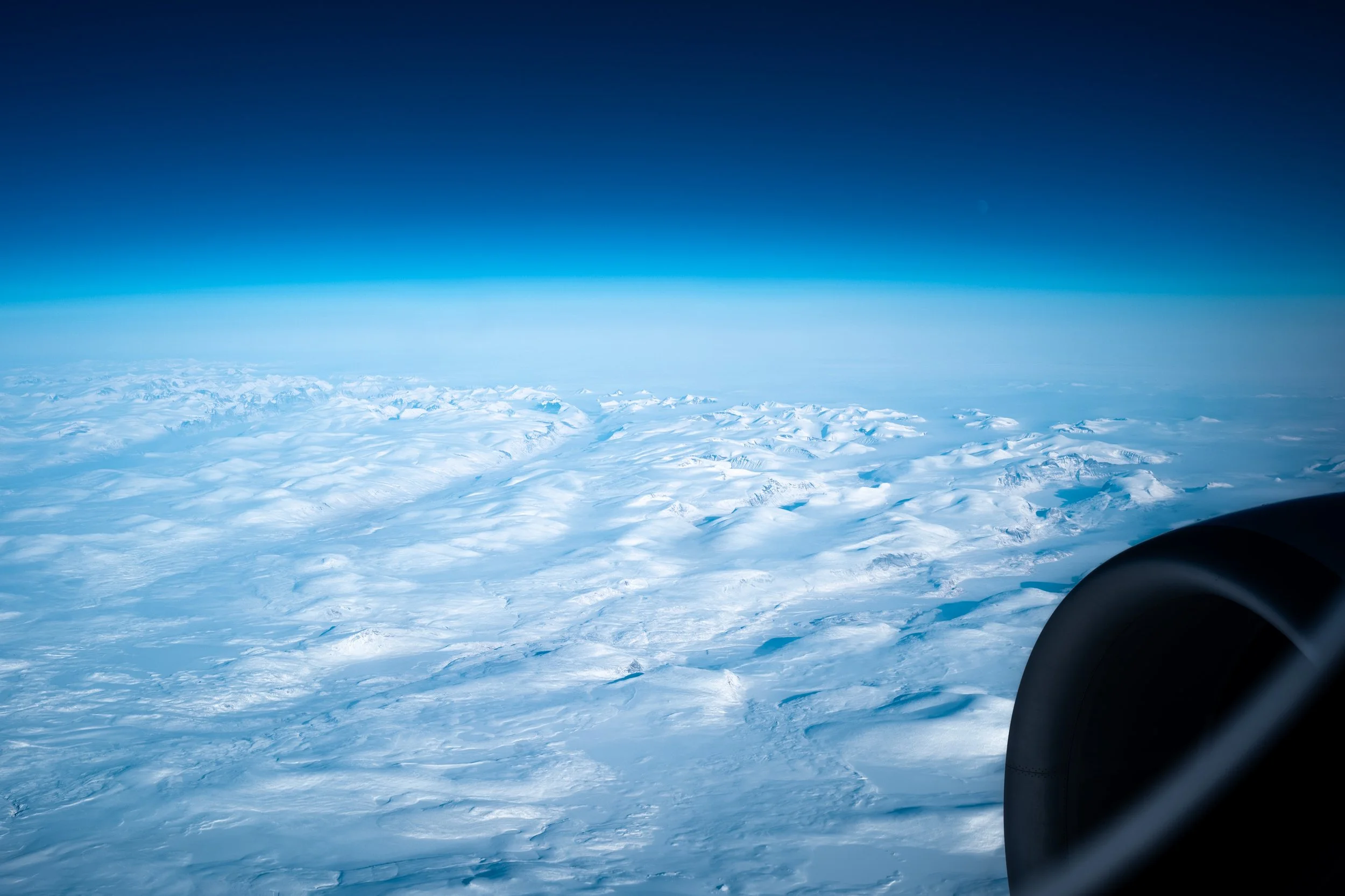 View of snow-covered mountains and icy terrain seen from an airplane window in flight, with part of the airplane engine visible on the right.