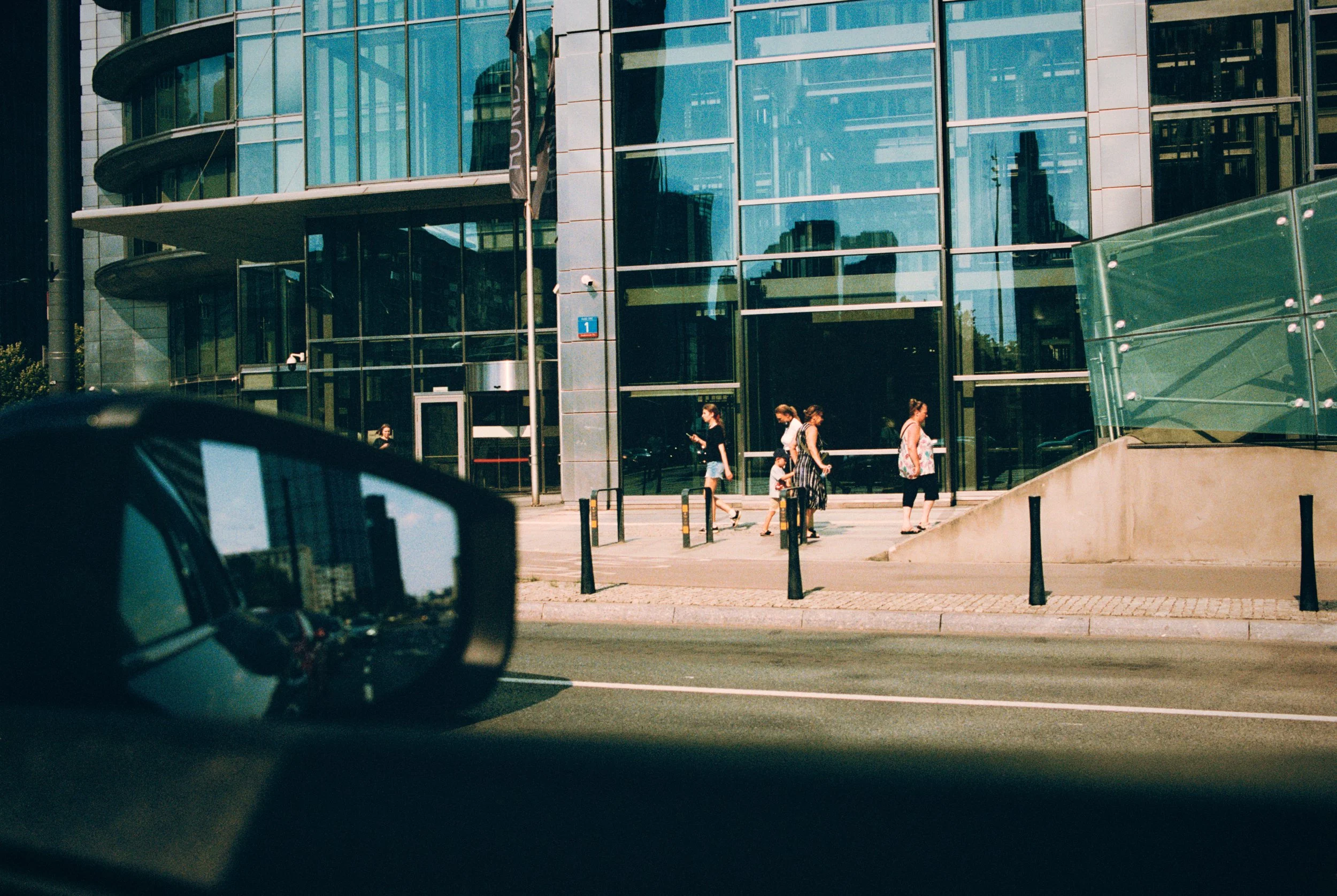 People walking on a city sidewalk in front of a modern glass building, with a car's side mirror visible in the foreground.