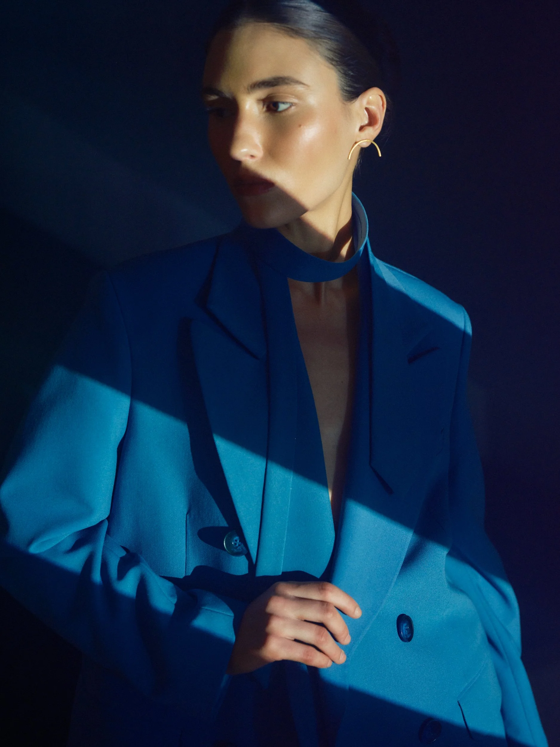 A woman wearing a blue suit with a high collar and gold earrings, standing against a dark background with dramatic lighting.