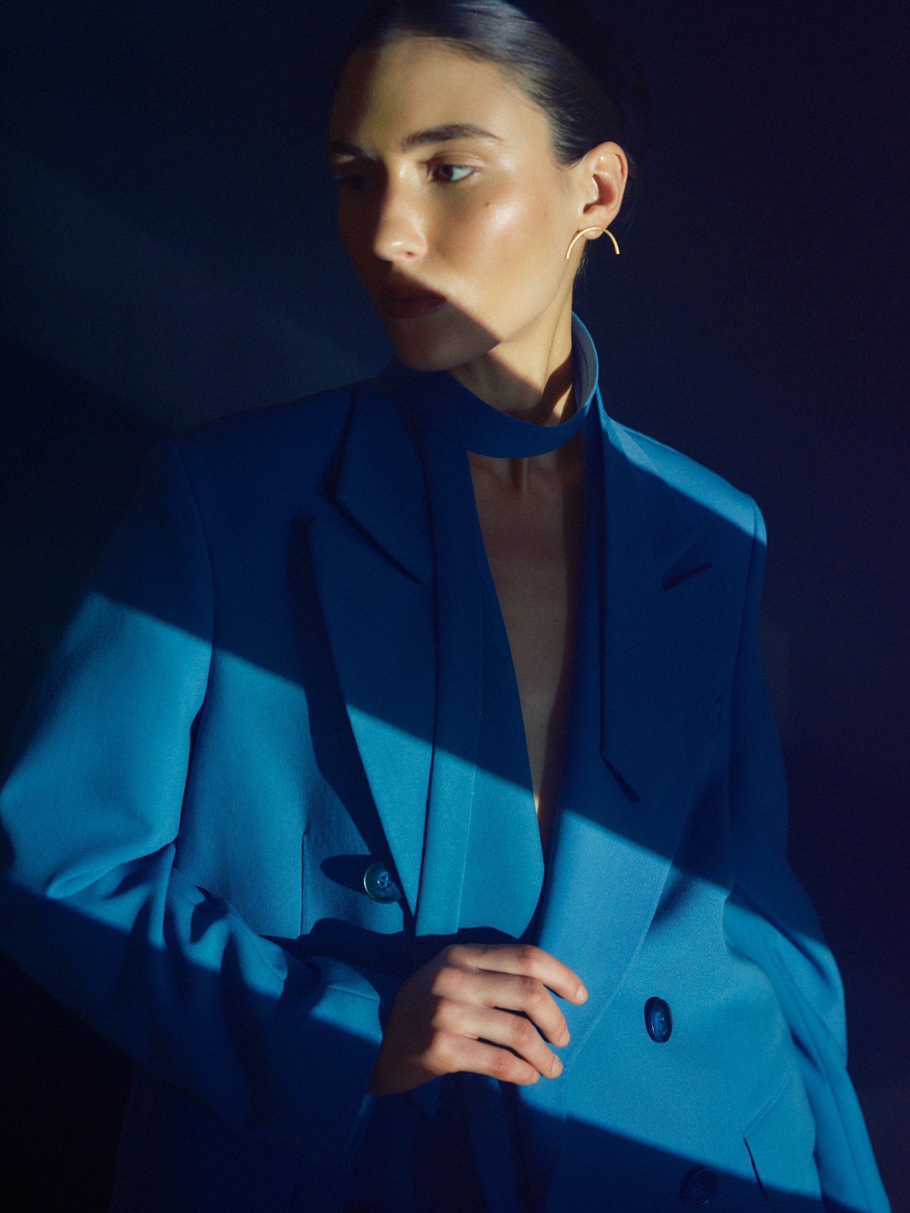 A woman wearing a blue suit with a high collar and gold earrings, standing against a dark background with dramatic lighting.