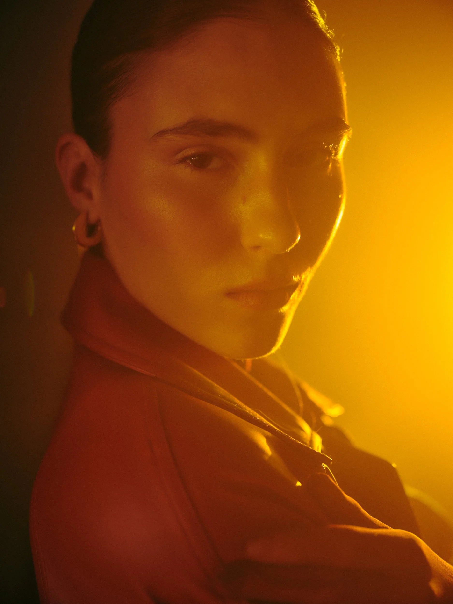 Close-up portrait of a woman with short dark hair and hoop earrings, illuminated by warm yellow-orange lighting, with a thoughtful expression.