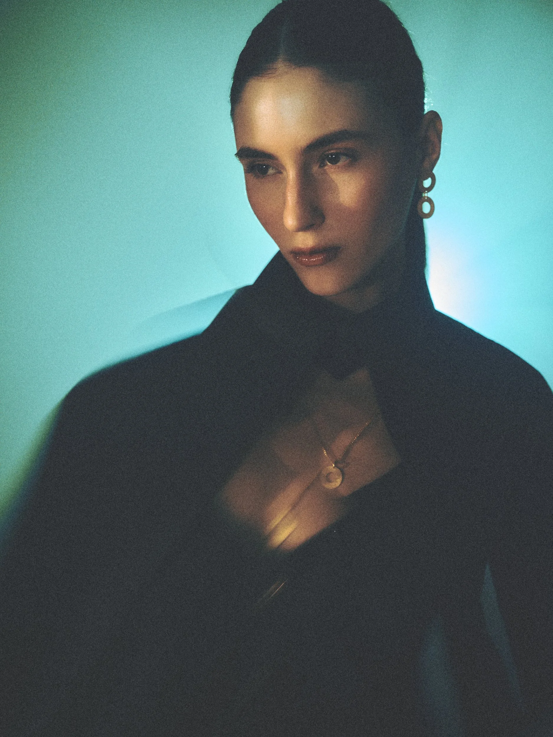 A woman with dark hair pulled back, wearing earrings and a black top, looking to her right with a neutral expression, against a greenish background with soft lighting.