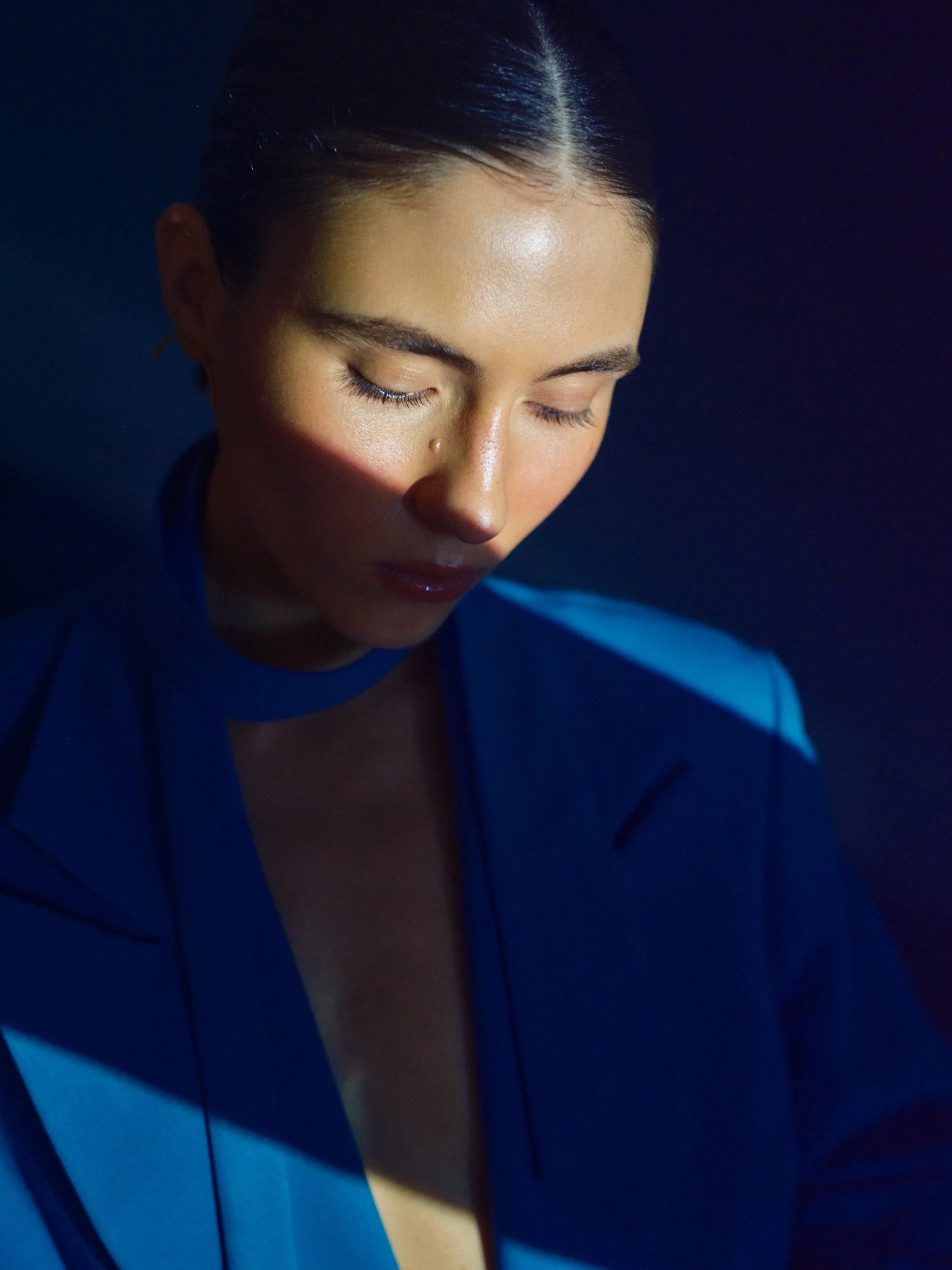 A close-up of a woman with closed eyes and dark hair tied back, wearing a dark top, against a dark background.