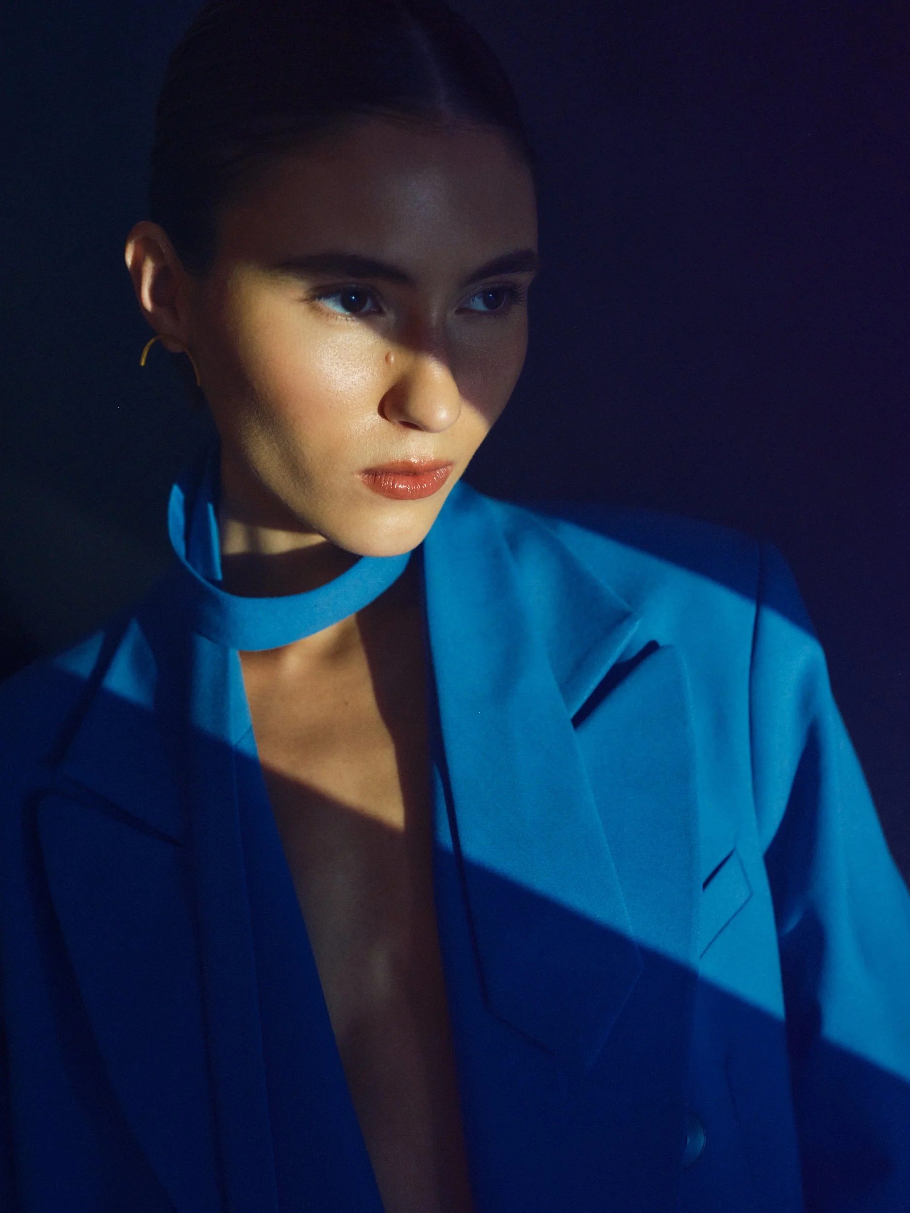 Close-up of a woman with dark hair wearing a blue blazer, gazing to the side, illuminated by a soft light against a dark background.