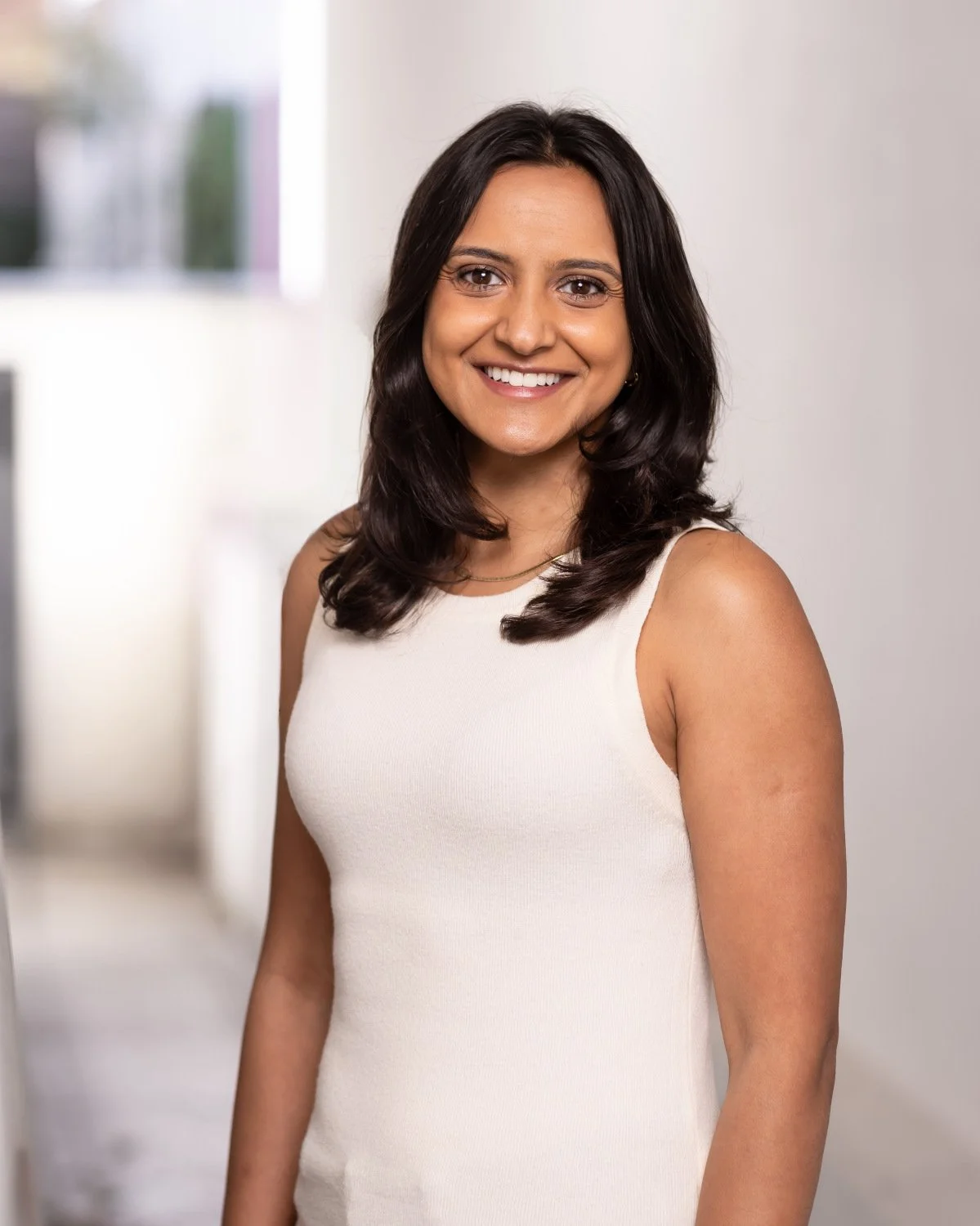  Summery outdoor headshot, set against a white walled building, of woman in a smart white vest and dark hair - with a friendly smile. 