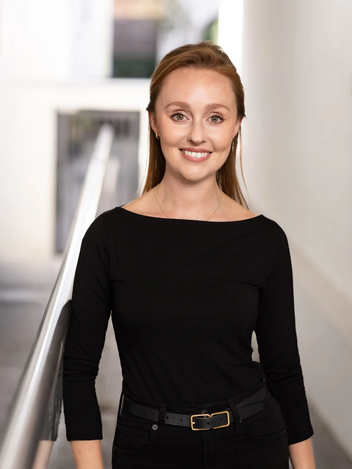   Portrait of a confident professional woman with long dark blonde hair and a fitted balck top, against a white exterior wall, with soft natural light highlighting her face, creating a warm and approachable headshot.  