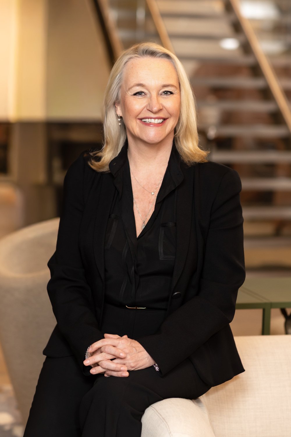  A blonde haired professional woman, dressed in a sharp black suit leans on an office sofa and smiles confidently. 