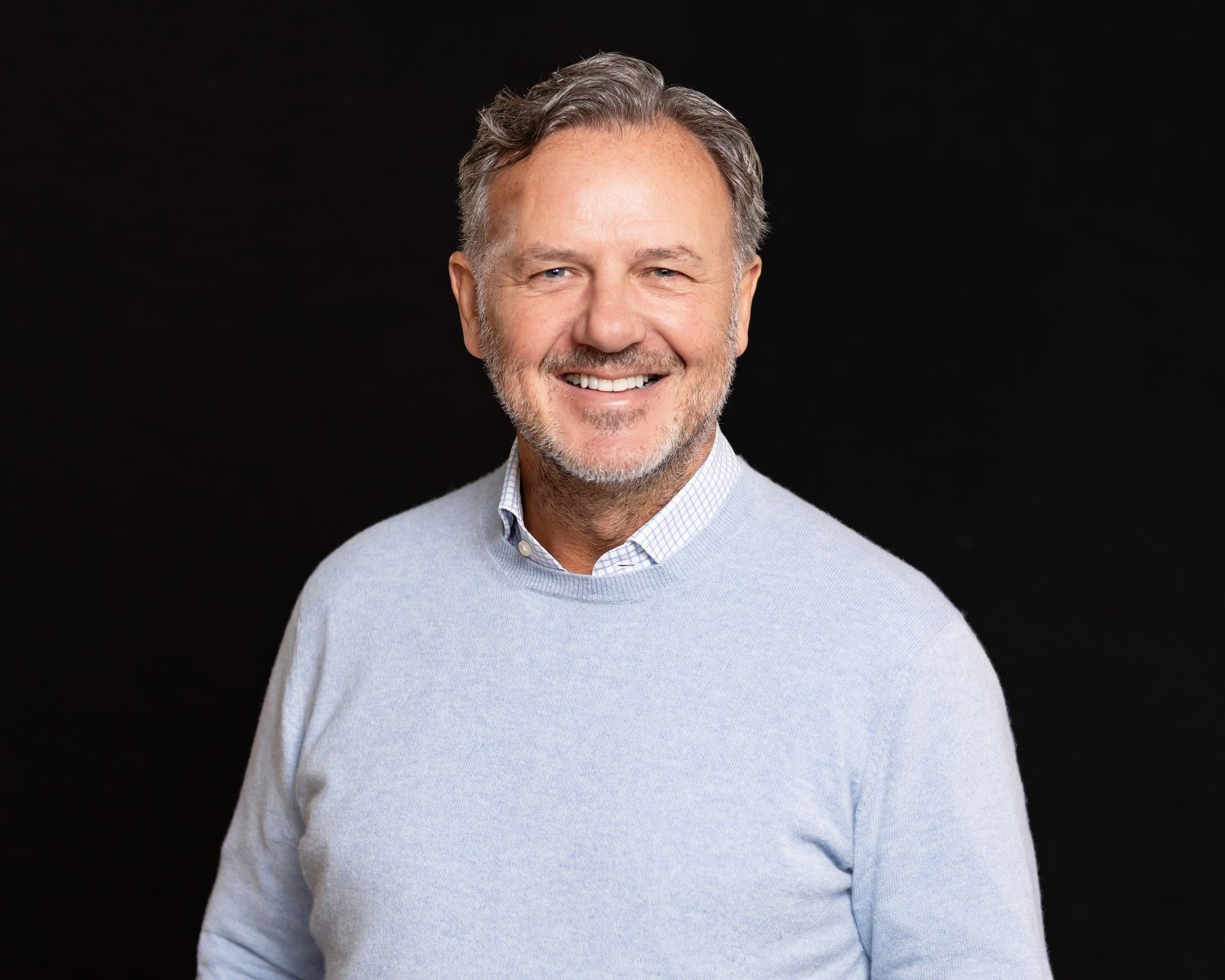  Grey haired man wearing a light blue jumper posing in front of a jet black background, with a warm friendly confident energy. 