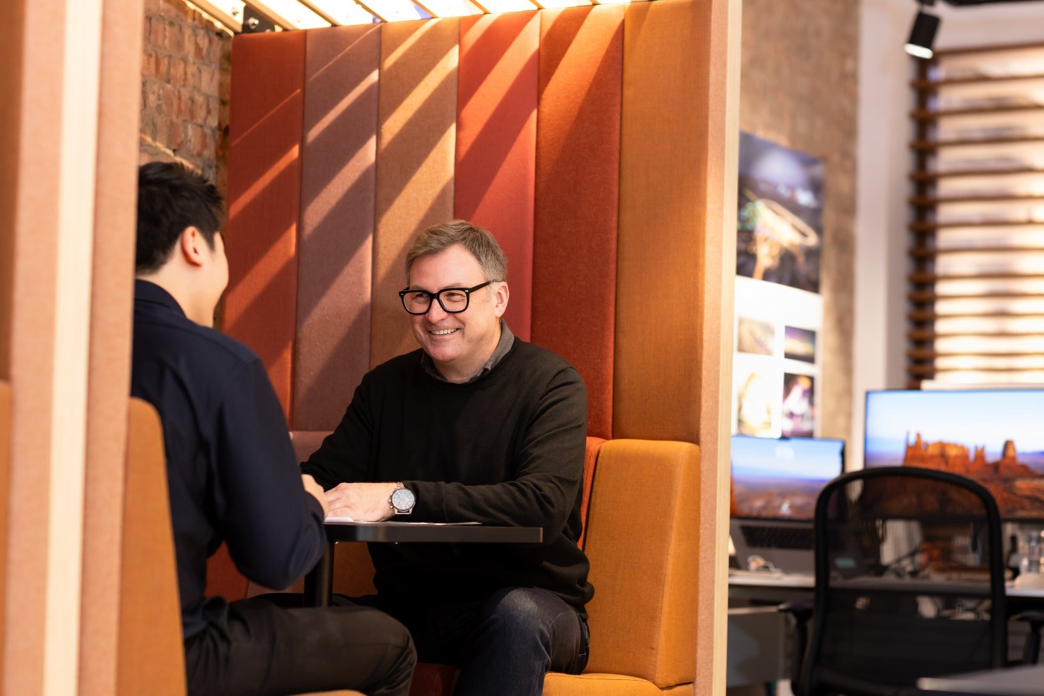  Two male creatives have a friendly  meeting in an orange workspace booth. Both men are dressed in black. 