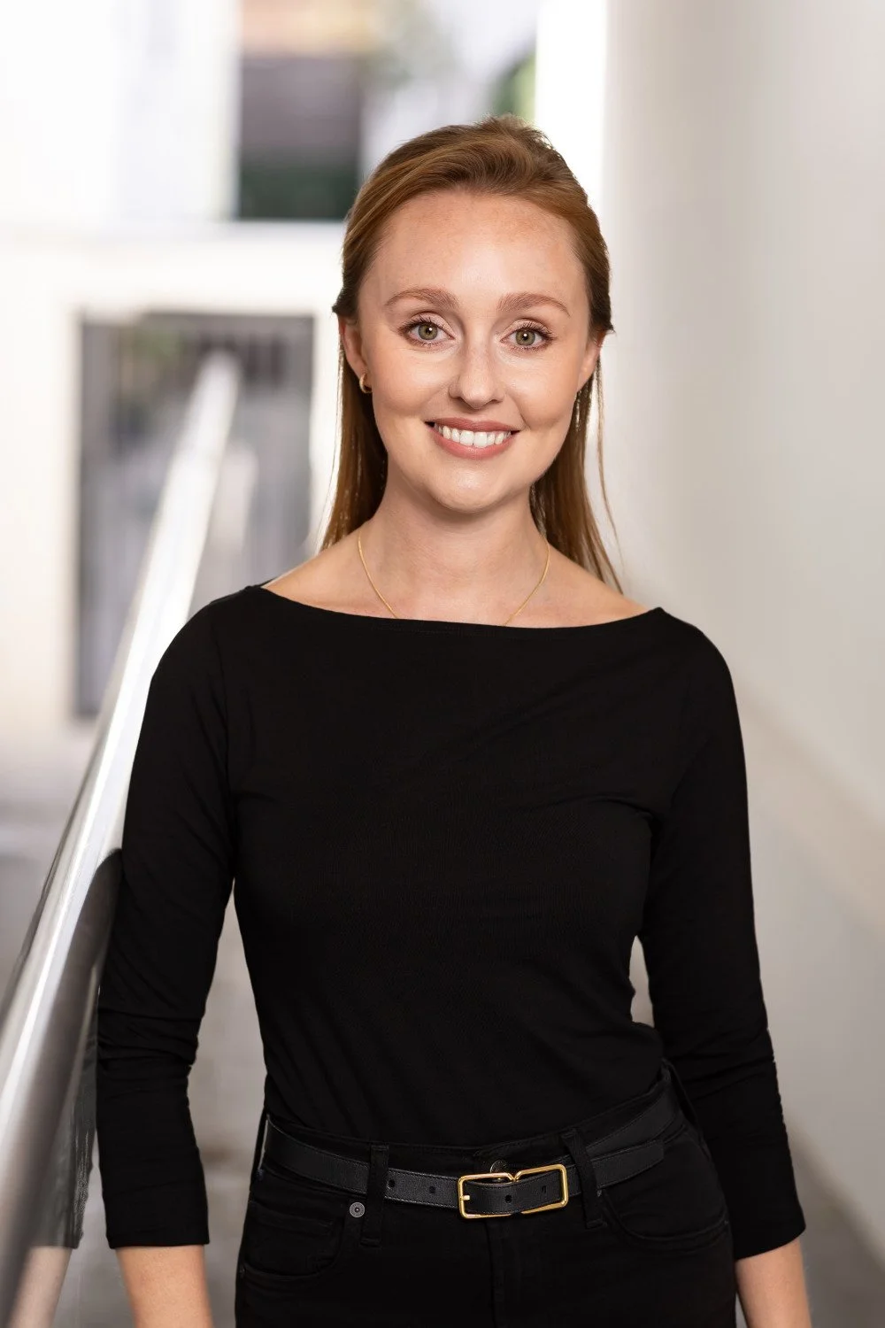   Portrait of a confident professional woman with long dark blonde hair and a fitted black top, against a white exterior wall, with soft natural light highlighting her face, creating a warm and approachable headshot.  