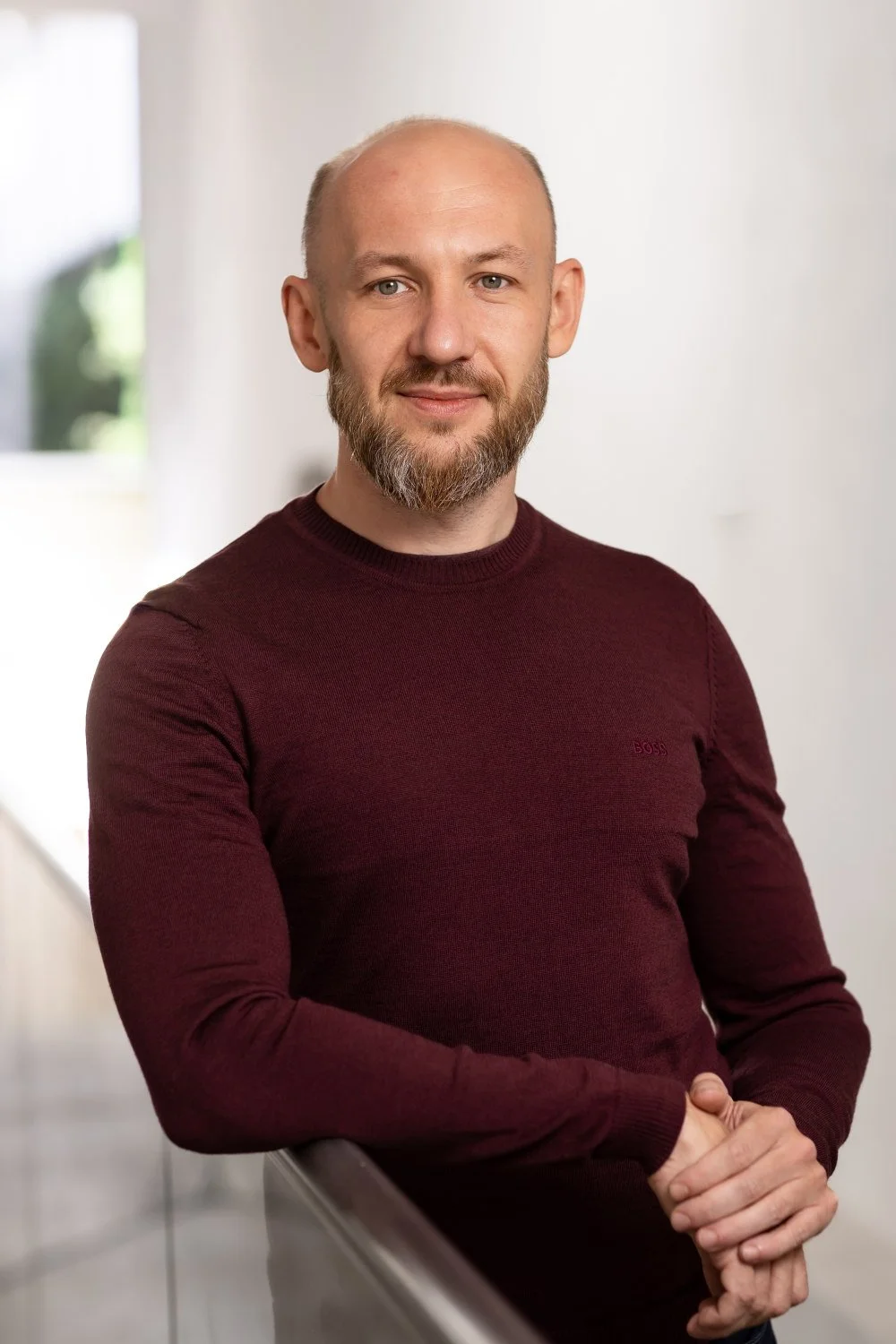  A professional man, with a beard and shaved head, leans on a glass wall. Photographed outside with a bright airy background, with flattering light and a confident calm expression. 
