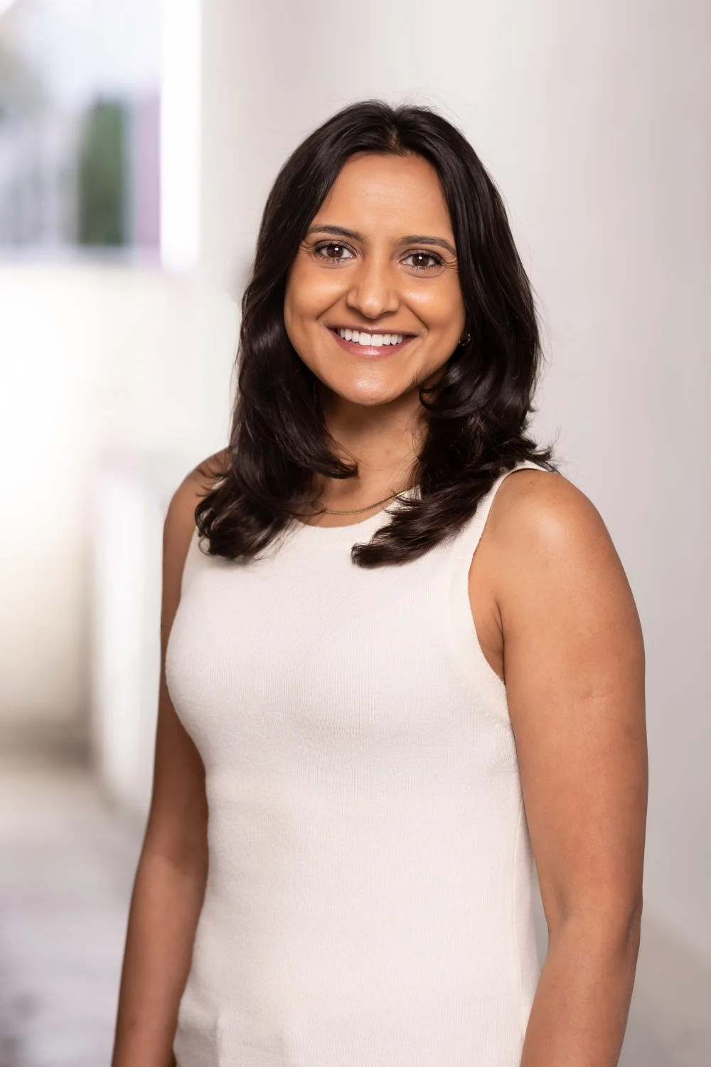  Summery outdoor headshot, set against a white walled building, of woman in a smart white vest and dark hair - with a friendly smile. 