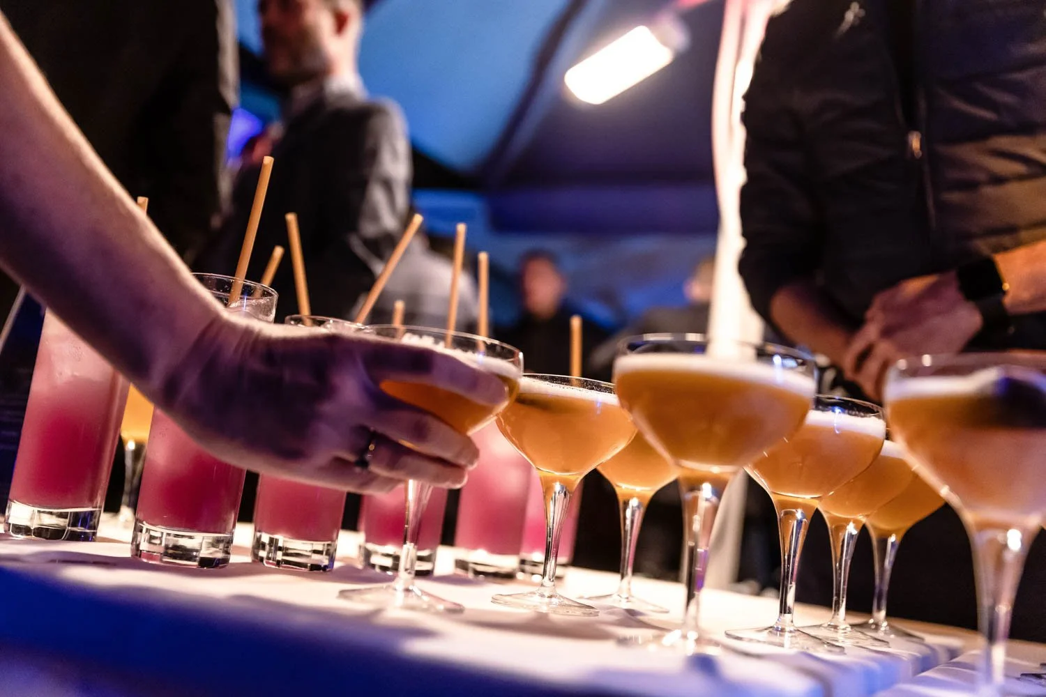  A hand reaches for a cocktail from a table of cocktails at an event 