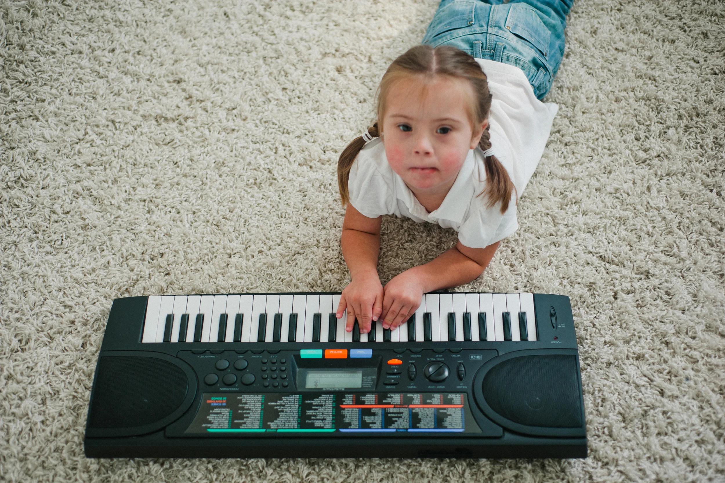 Young child with braided hair lying on a carpet playing an electronic keyboard.