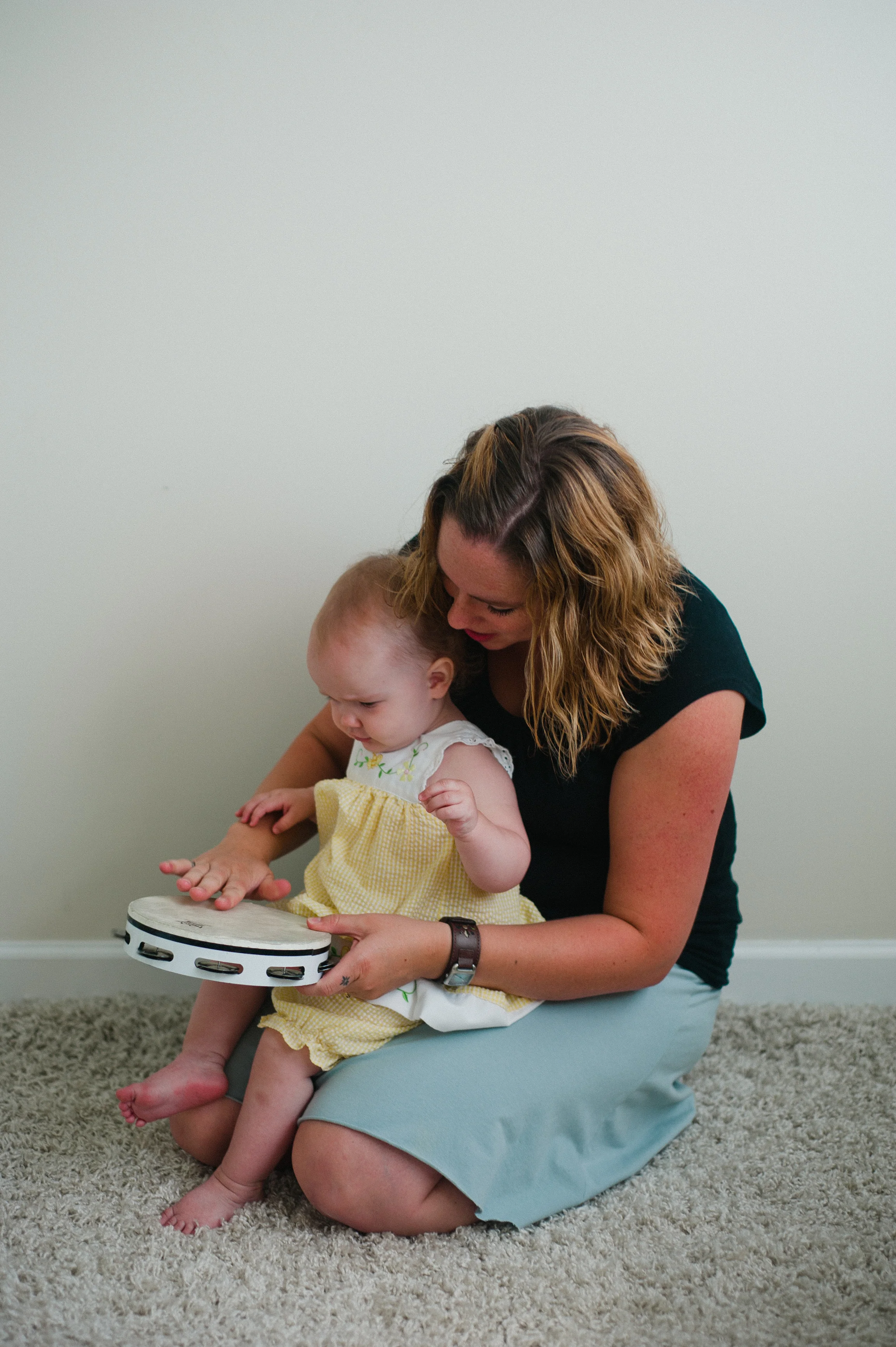 Woman sitting on floor holding baby playing tambourine.