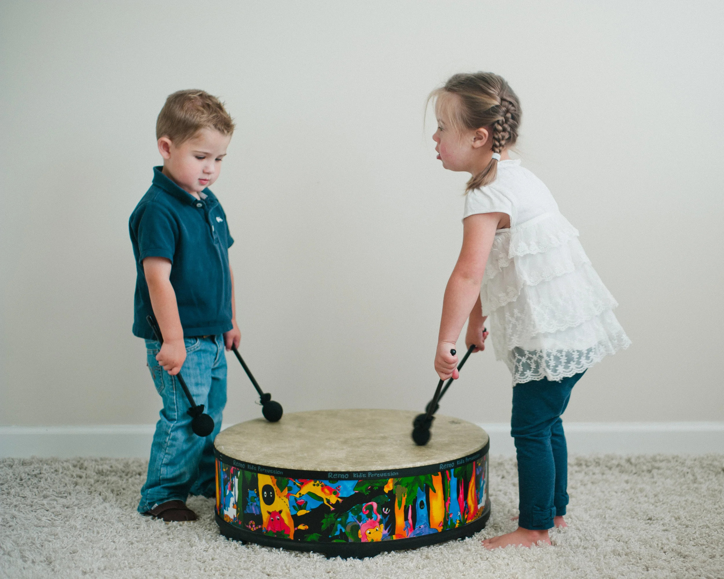 Two children playing a colorful drum on a carpeted floor, each holding a mallet.