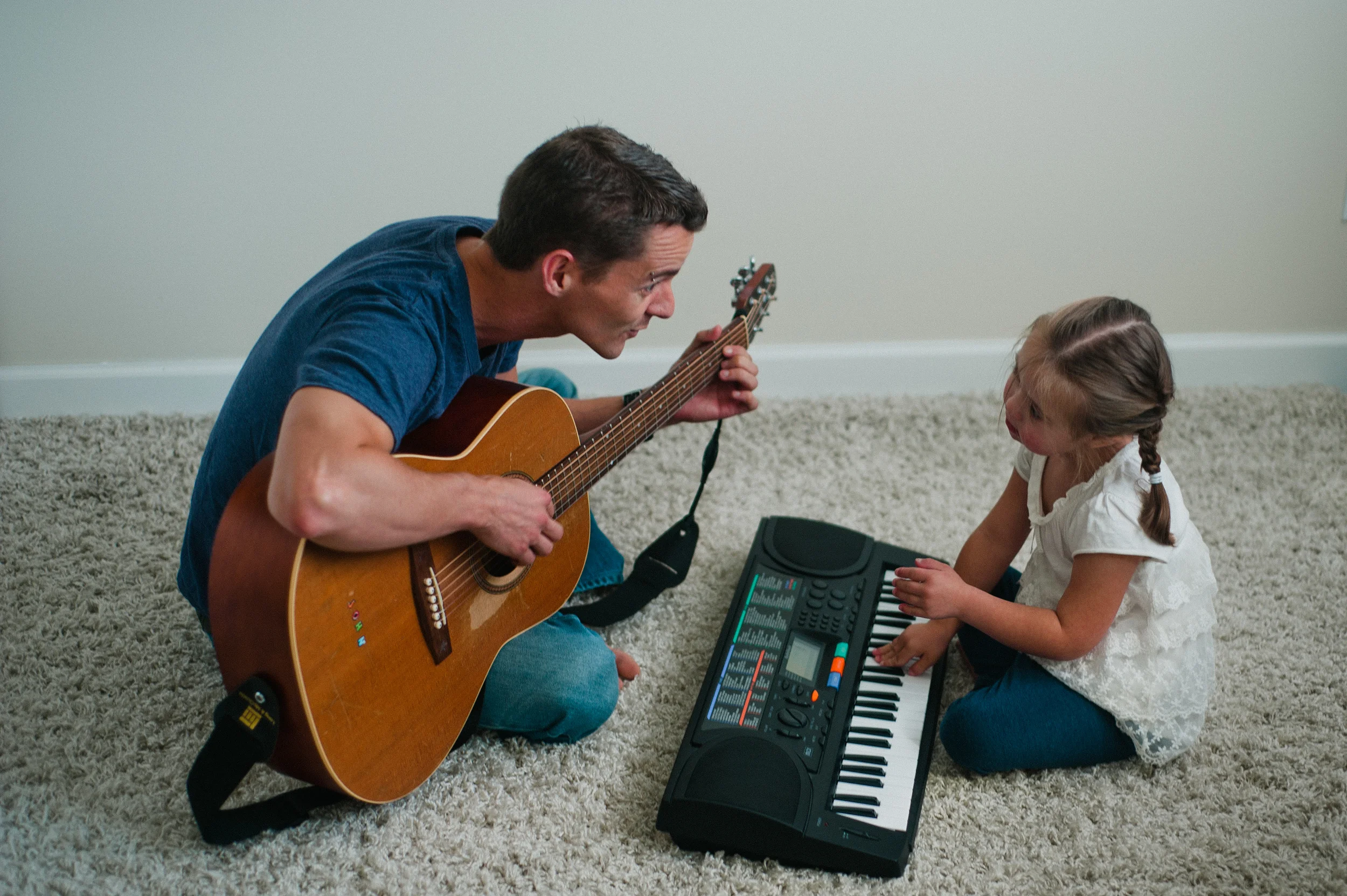 Man playing an acoustic guitar and a child playing a keyboard on a carpeted floor.