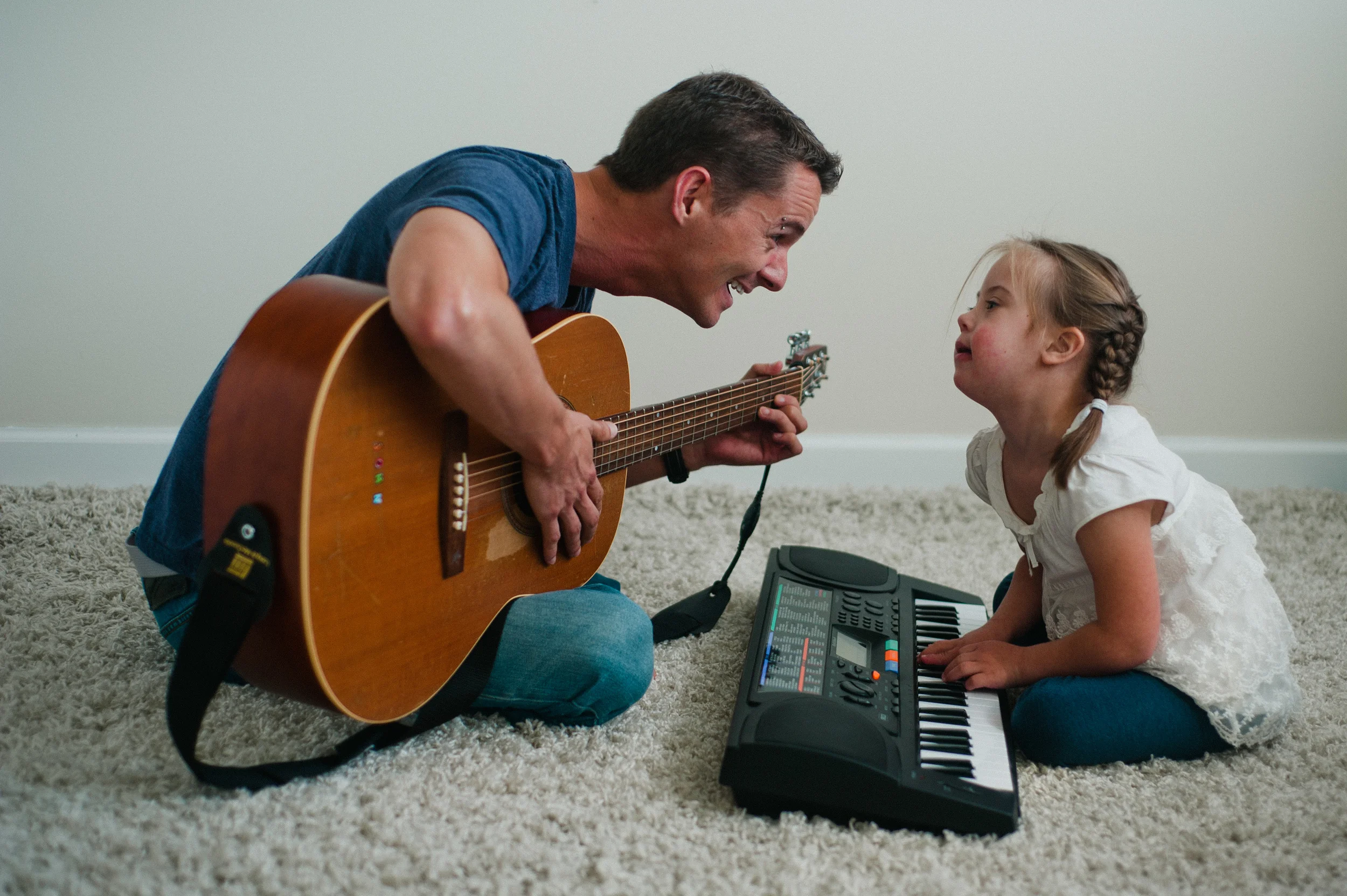A man playing guitar and a young girl playing a keyboard, both sitting on a carpeted floor, smiling and interacting.