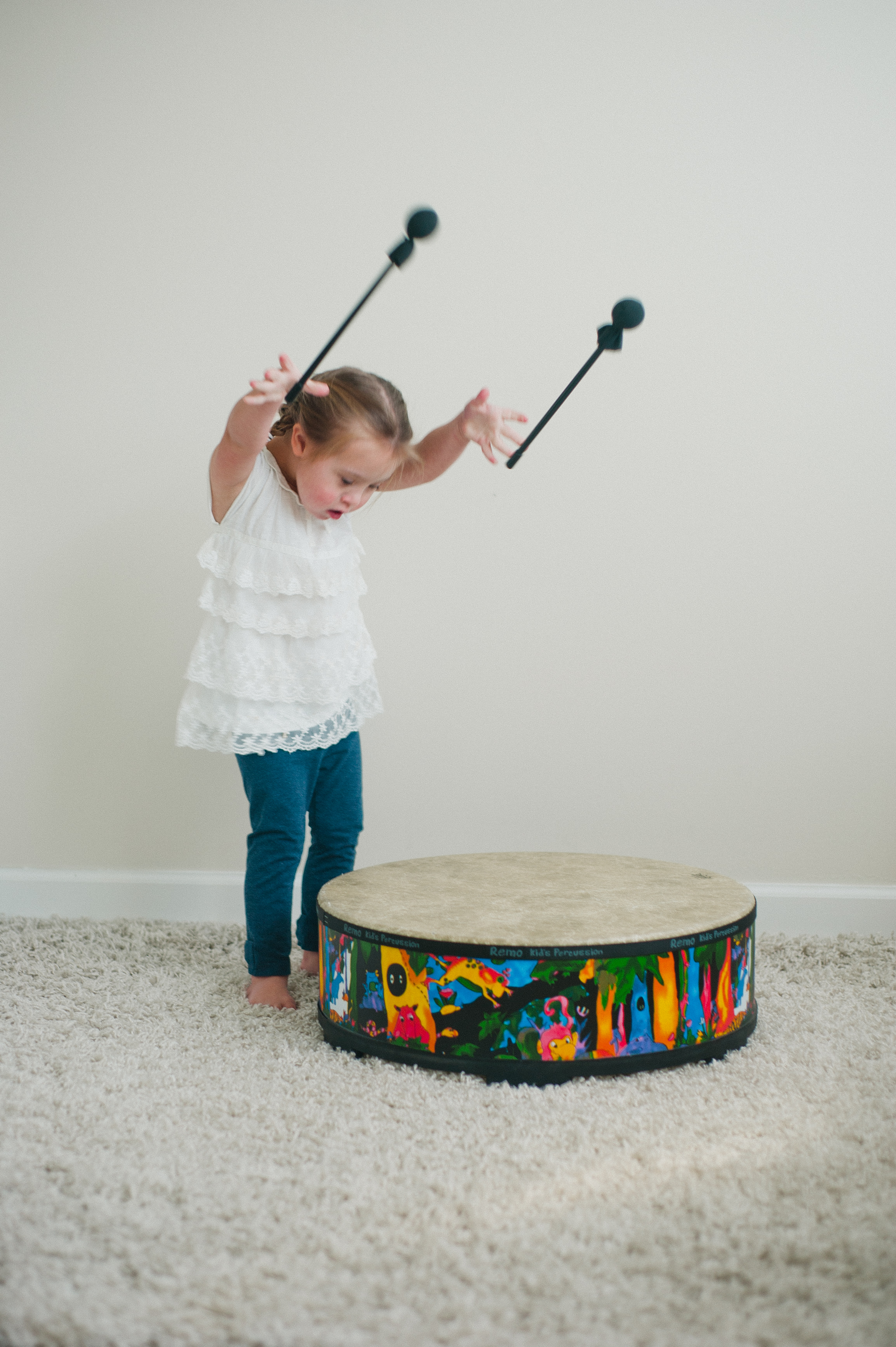 Child playing with a colorful drum using mallets on a carpeted floor.