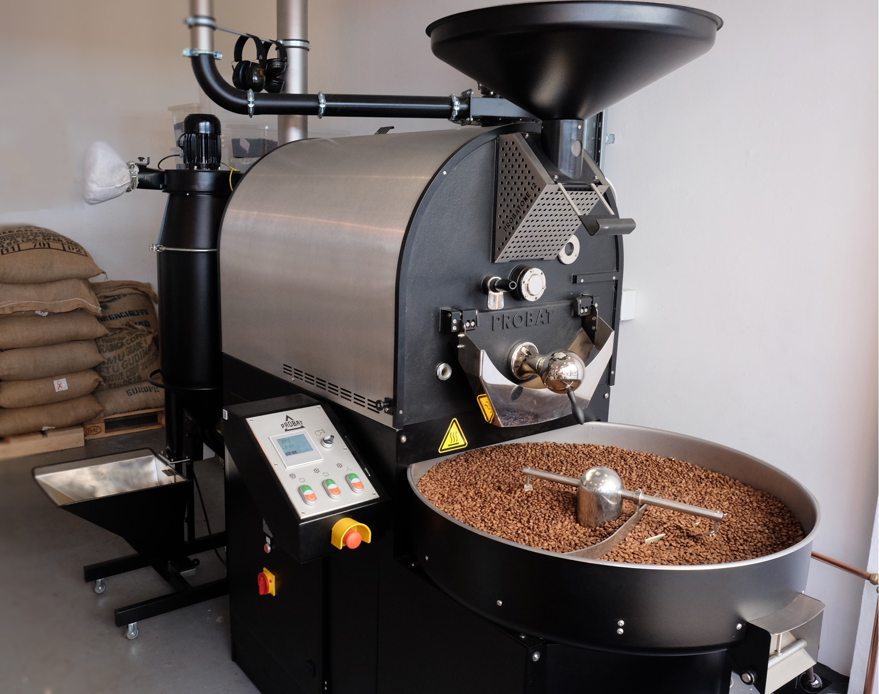 A commercial coffee roasting machine with a digital control panel, a large roasting drum, and a cooling tray filled with roasted coffee beans, situated in a coffee roasting facility.