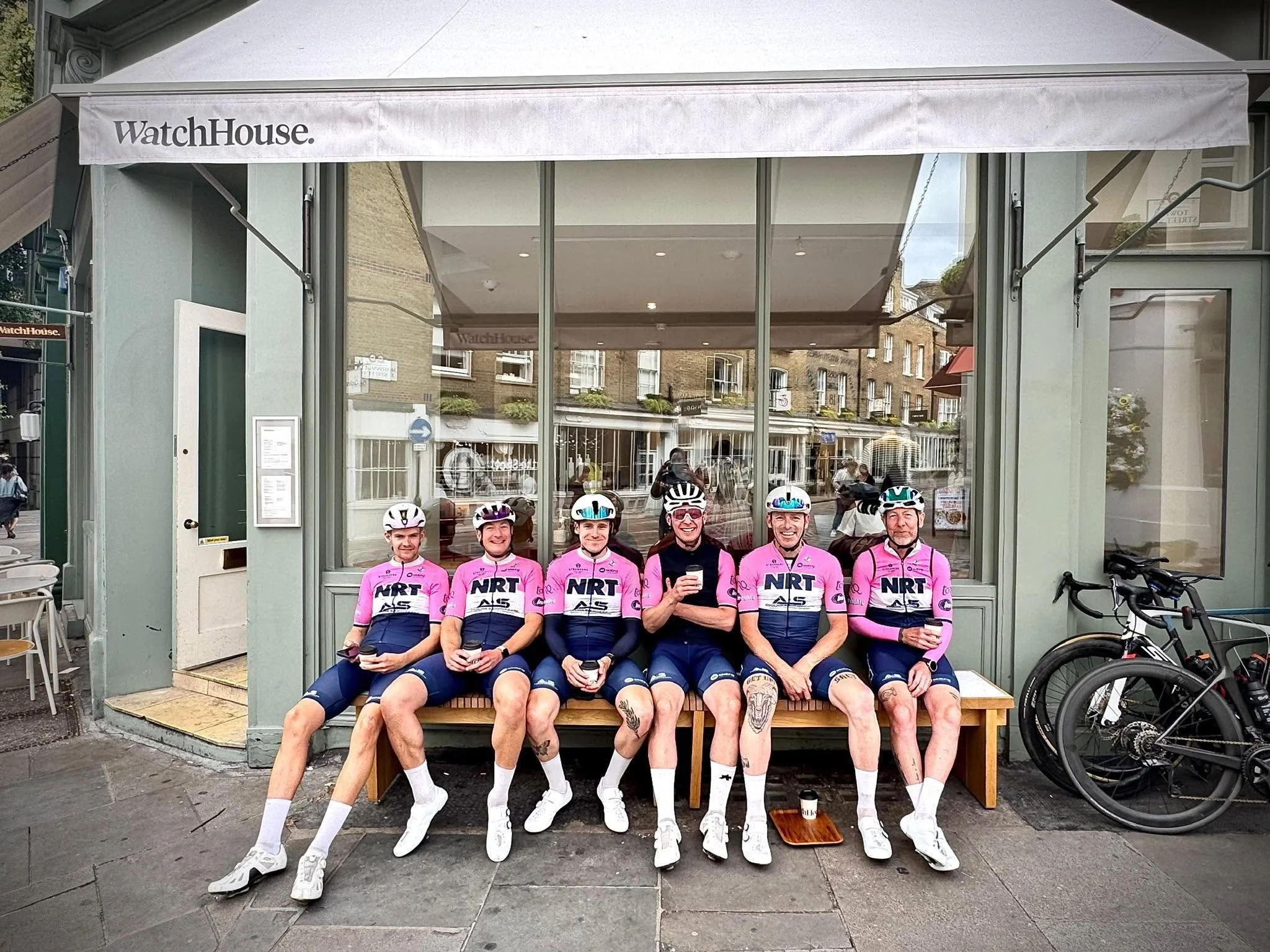 Six cyclists in pink and navy uniforms sitting on a bench outside a storefront, holding drinks. There are helmets on their laps and some tattoos visible on their legs. They are smiling and posing for a photo.