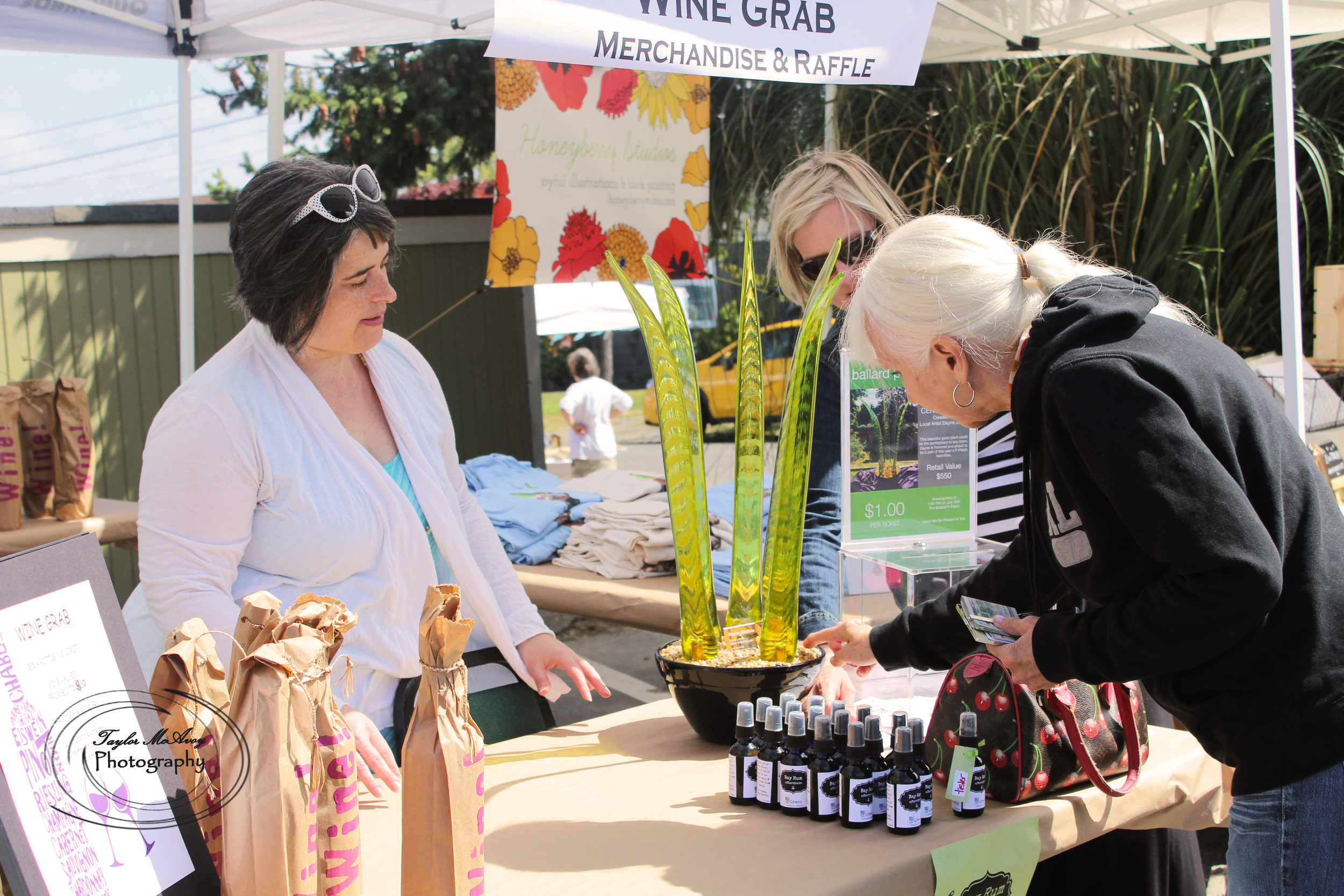  Dahlia Cohen chats with customers about the Wine Grab as they sign up for a raffle for a piece of glass art by Dayne Lopez. The Wine Grab features volunteer donated wine organized in Red or White and the rest is a mystery until unwraped.&nbsp; 