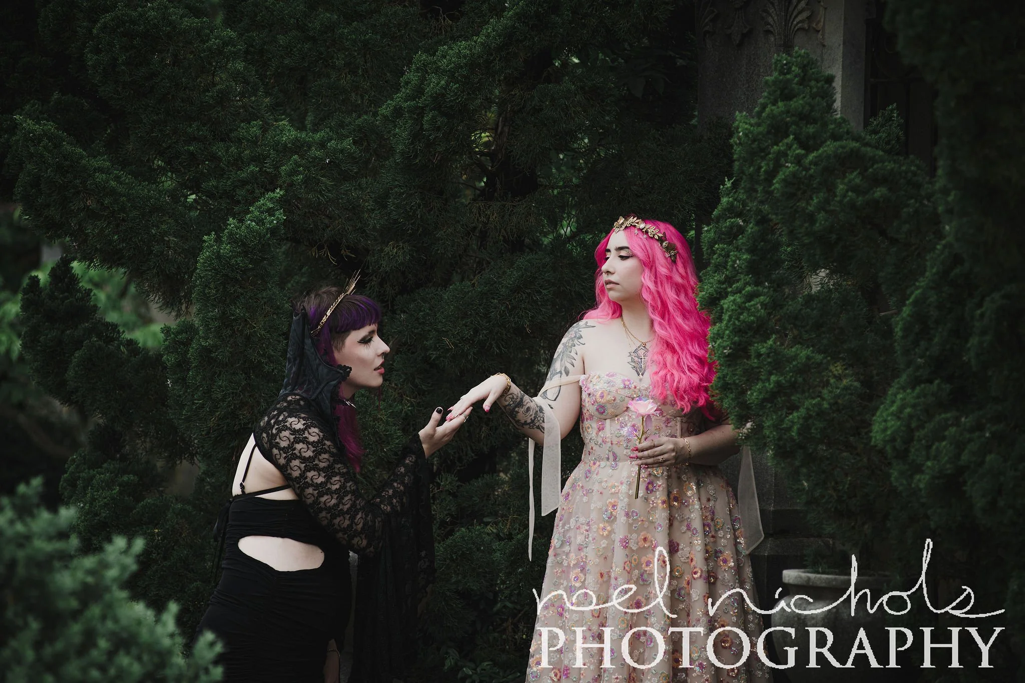 Two women with colorful hair, one with pink and one with purple, in a lush garden with cypress trees. The scene is dark and romantic.