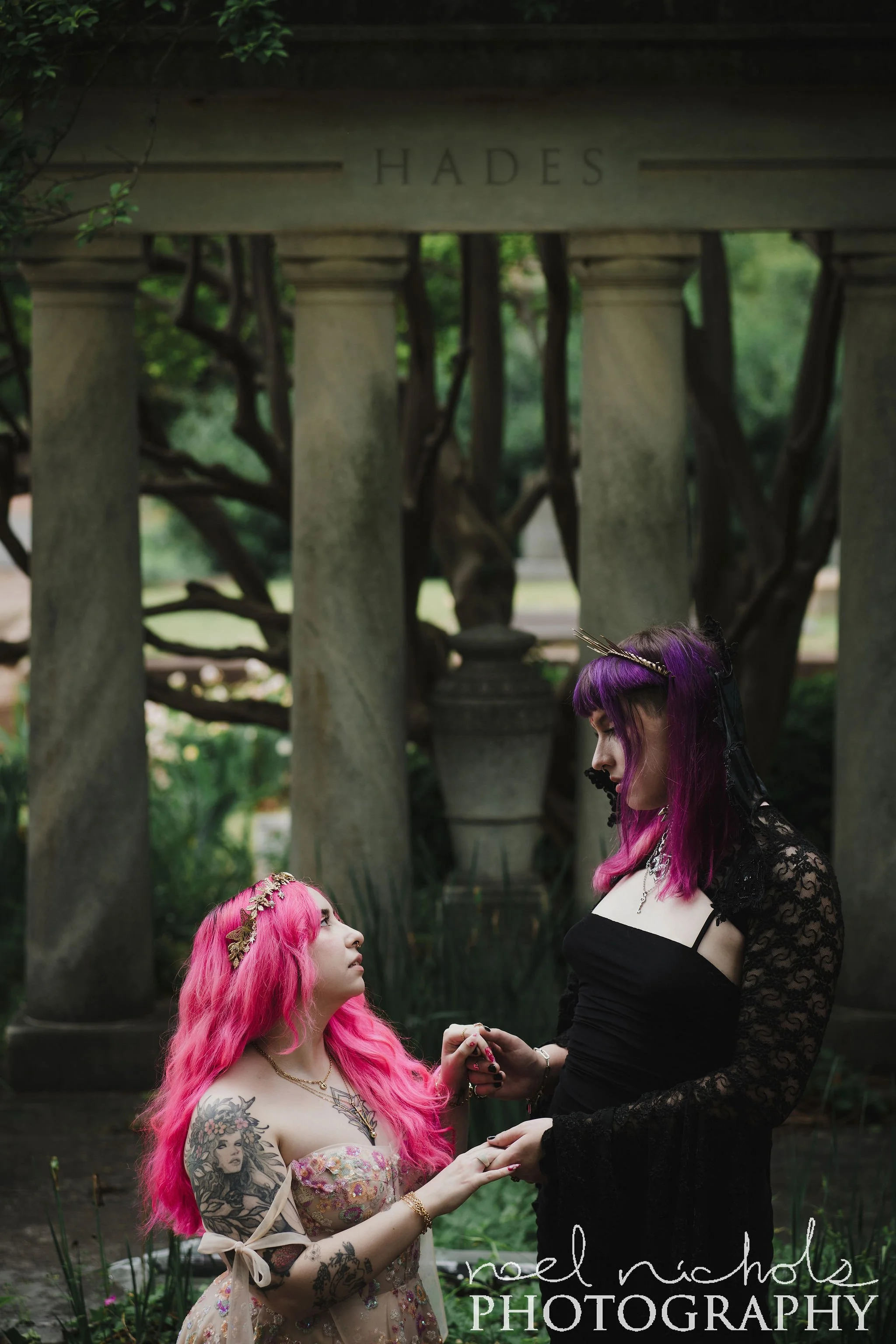 Two women with colorful hair, one with pink and one with purple, in a lush garden with a columned structure. The scene is dark and romantic.