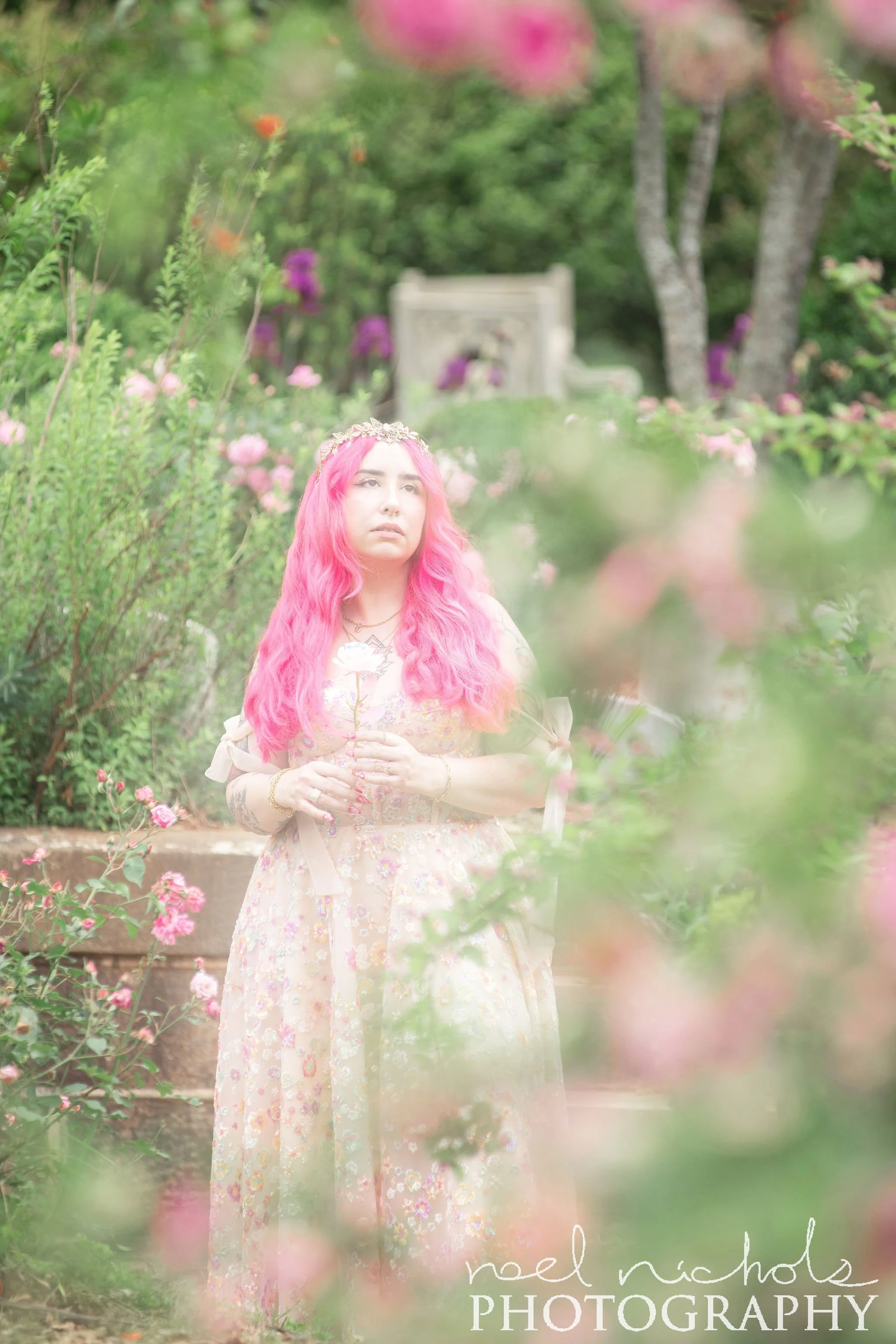A photo of a woman with long vibrant pink hair in a lush garden. The garden has pink and white blooms. 