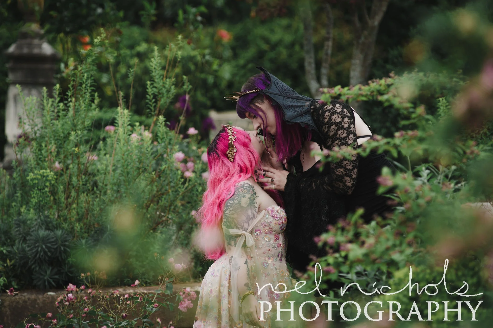 Two women kissing. They have colorful hair, one with pink and one with purple, in a lush garden with pink and purple flowers. The scene is vibrant and romantic.