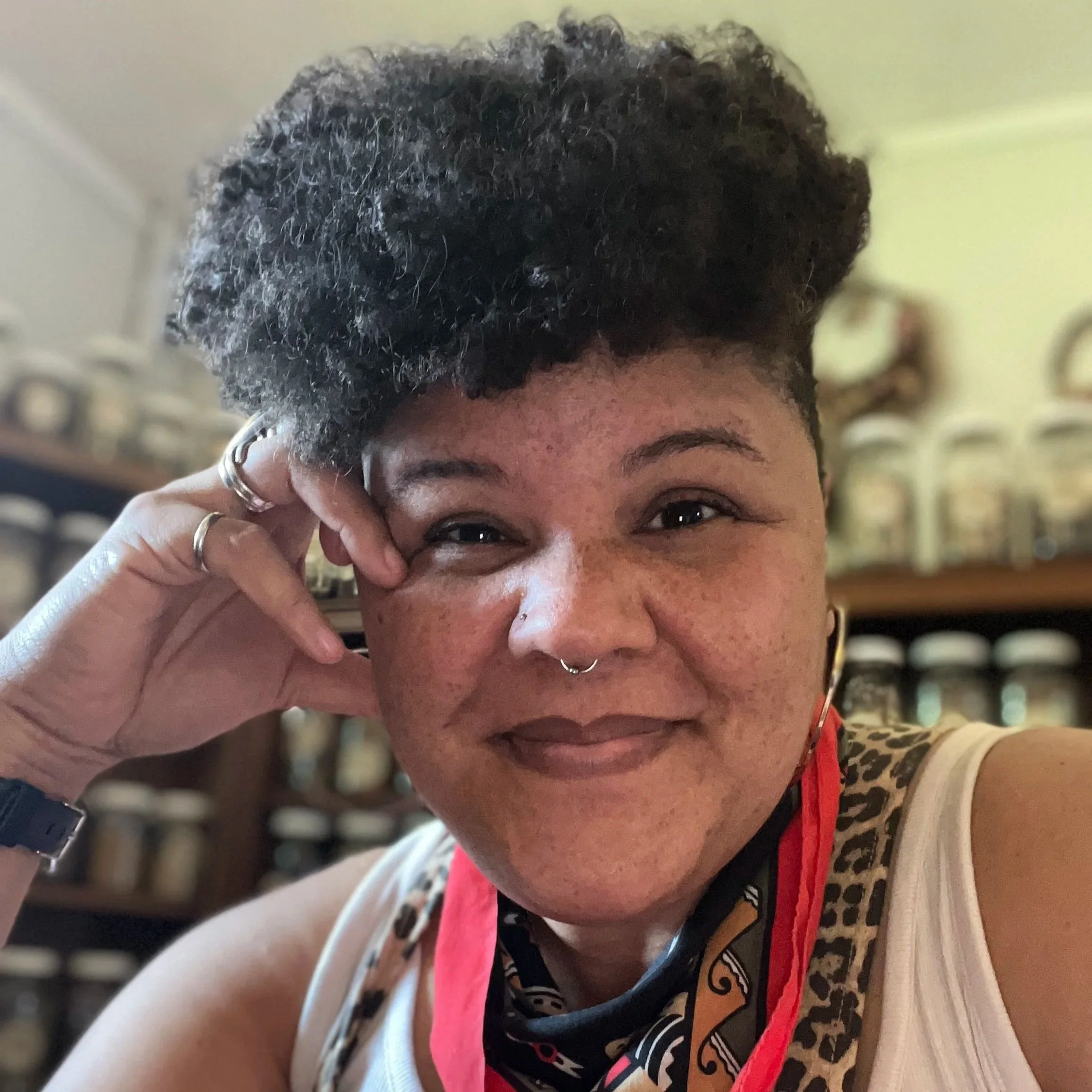 a closeup of a Black femme person with short natural hair, a septum ring, multiple rings and a watch (hand resting against temple), wearing a white tank top and a multi-colored neckerchief, against a blurred background of a wall of jarred herbs