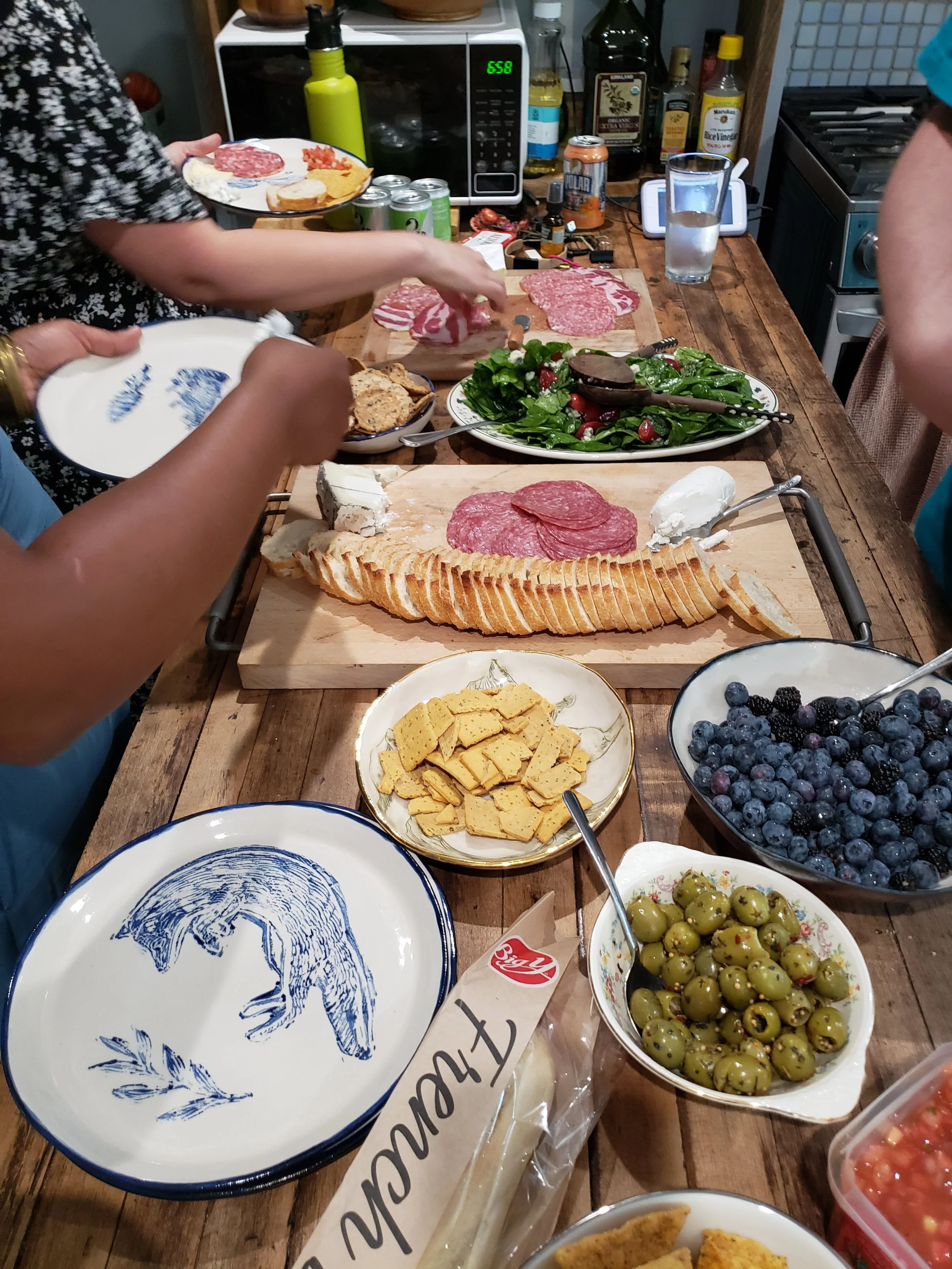 An image of food in dishes spread across a wooden surface: crackers, meats, berries, cheeses, salad, bread, olives; a white stoneware plate with a painted fox and sprig of herb in blue; three arms seen reaching and serving food