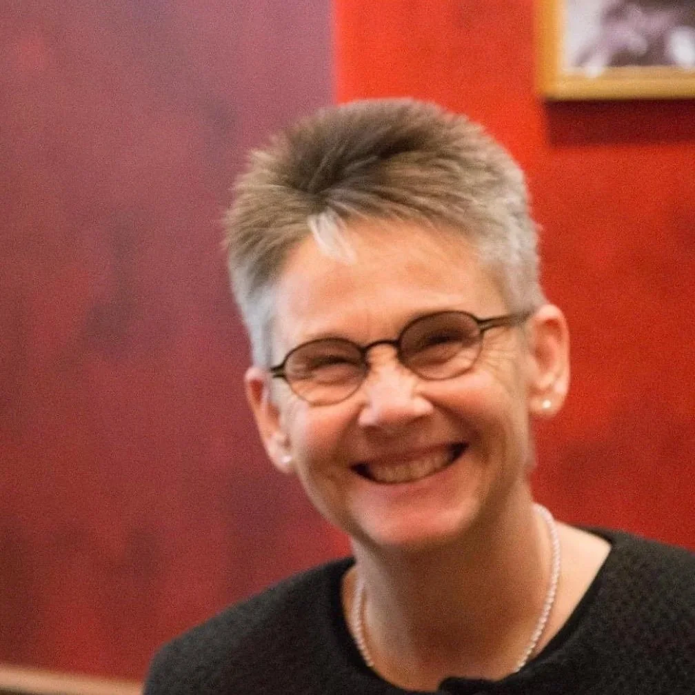 a closeup of an older white woman with short grey hair, round dark-framed glasses, a black top, pale stud earrings, and a short white beaded necklace, against a blurred background of a room with red walls and a framed painting