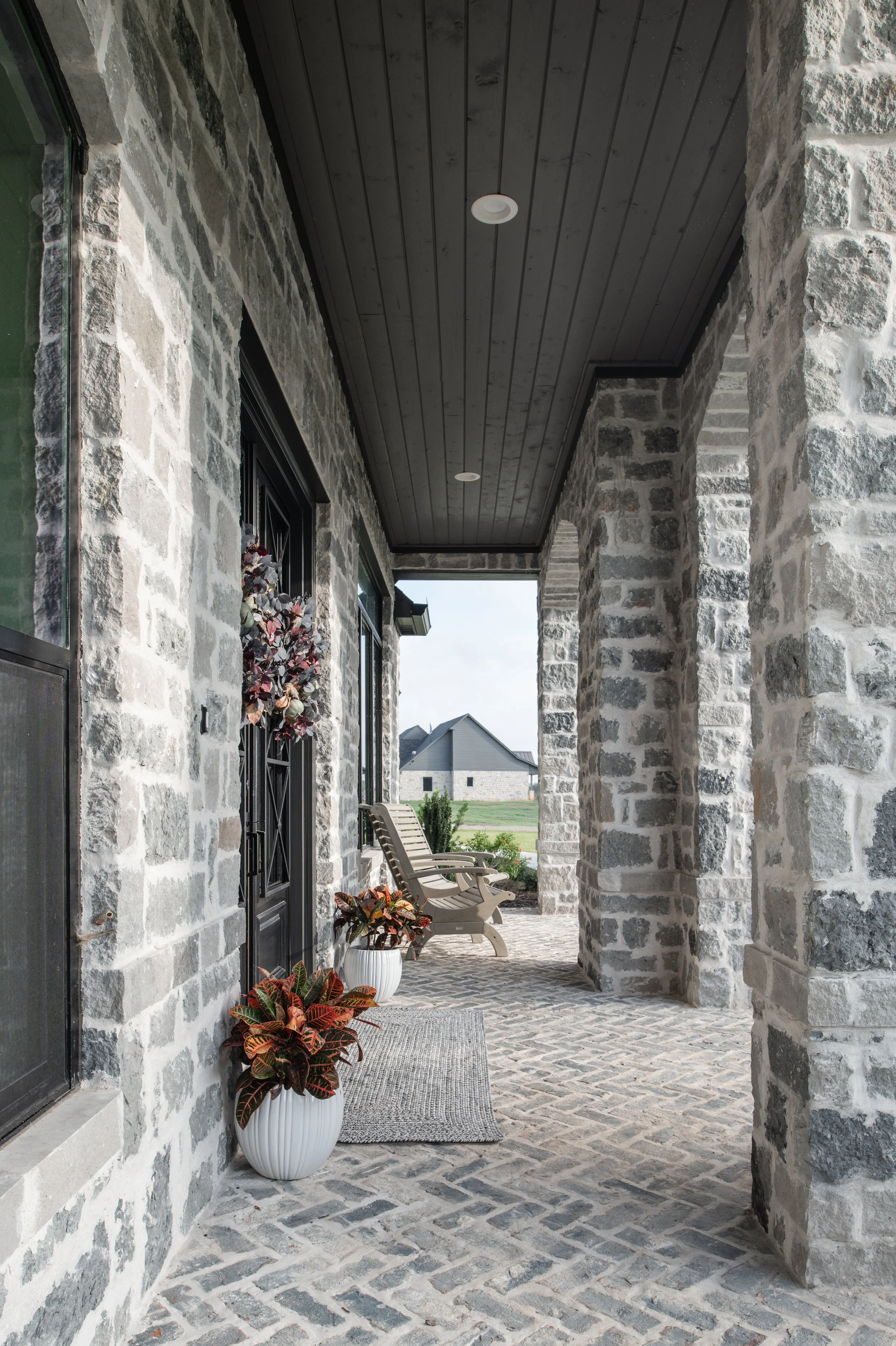 Front porch of a stone house with potted plants, chairs, and a doormat, overlooking a farm landscape.