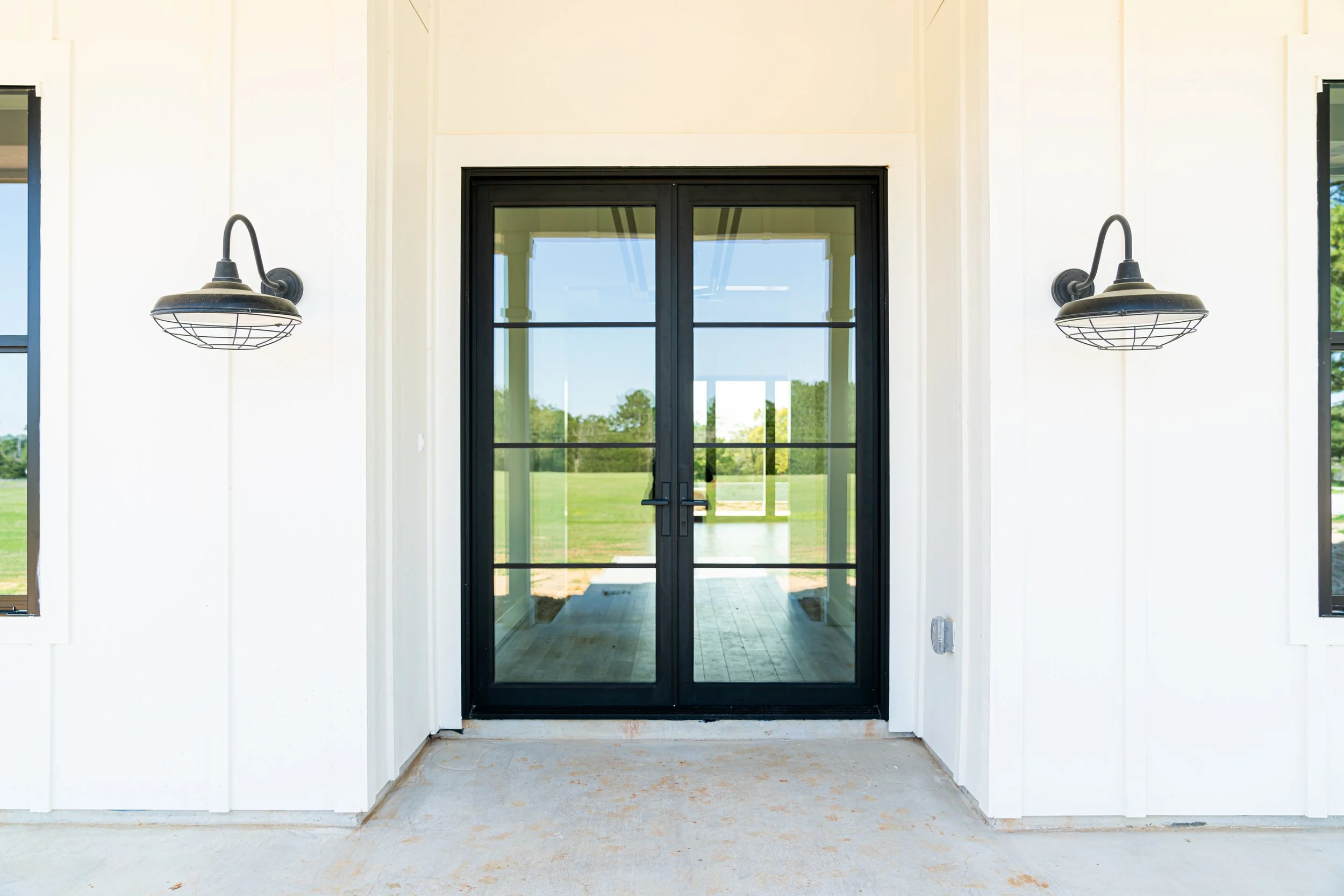 Front porch area with black-framed glass double doors, white walls, two black outdoor wall sconces, and a view of grass and trees outside.