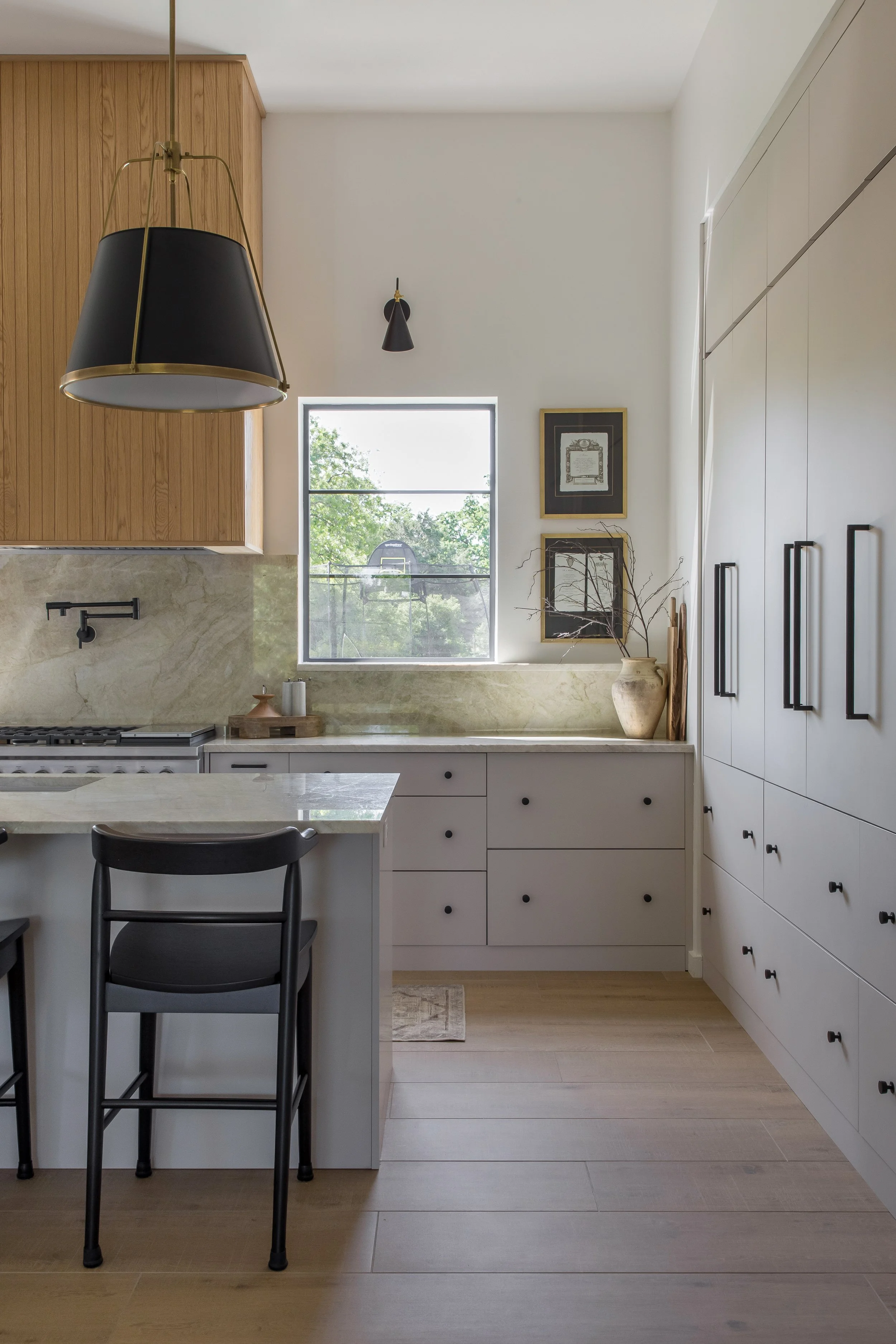 Modern kitchen with white cabinets, black handles, marble countertops, a window with trees outside, wooden ceiling cabinets, black hanging lamps, and framed art on the wall.
