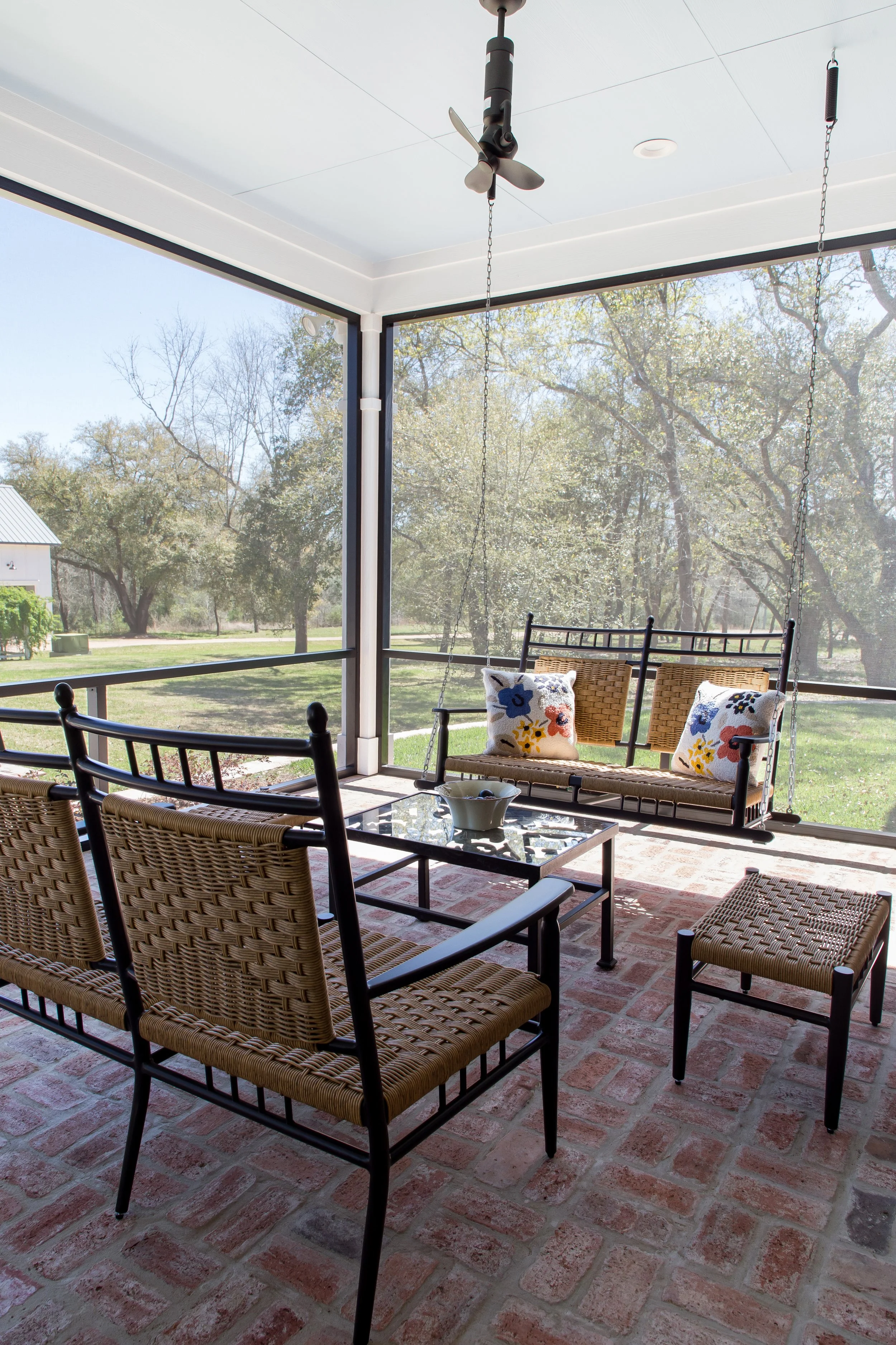 A screened porch with wicker and metal furniture, including a swing with floral cushions and a glass-topped coffee table, overlooking a backyard with trees and a house.