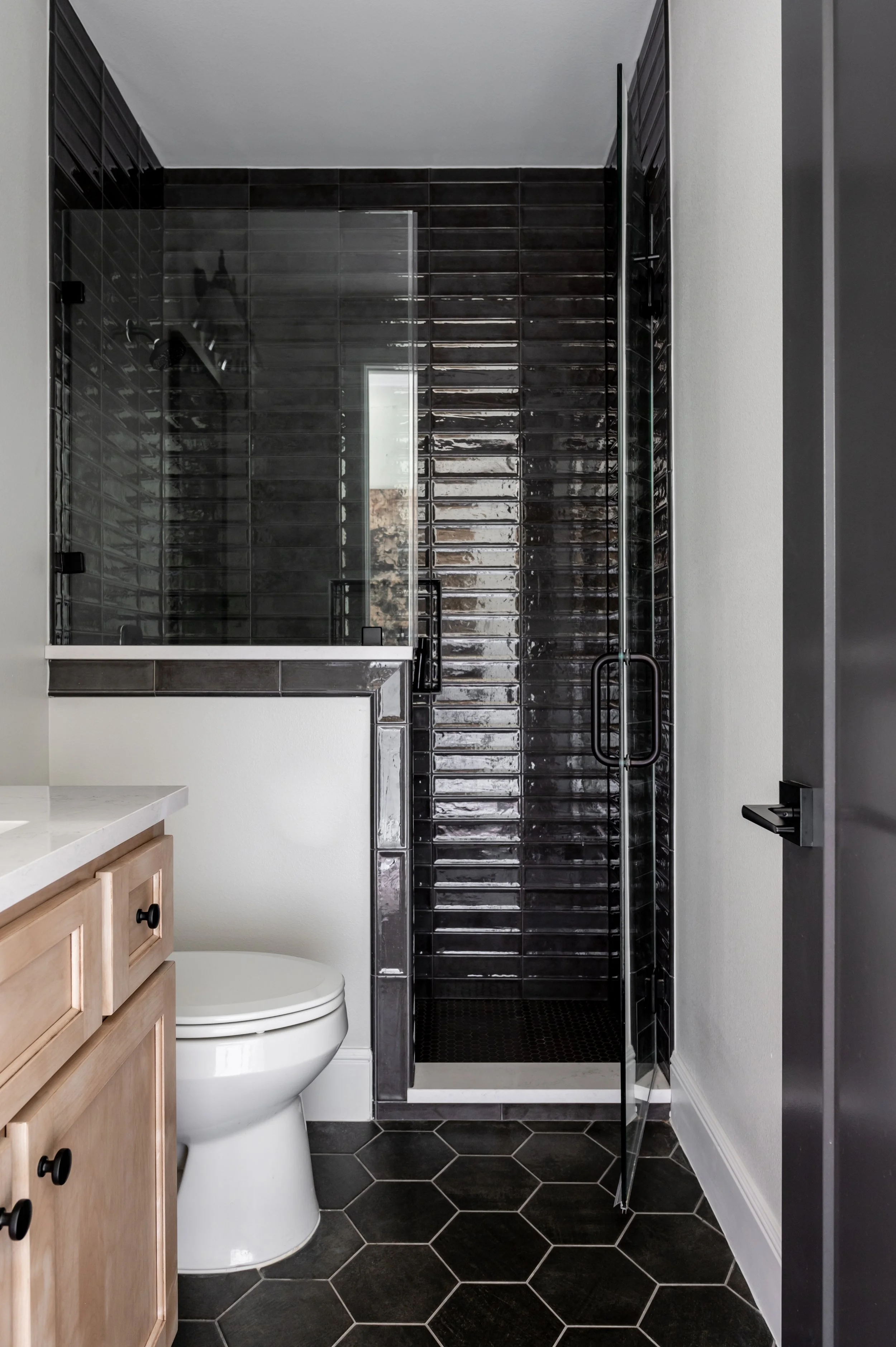 Bathroom with black tiled shower, a white toilet, black and wood cabinetry, black hexagon floor tiles, and a gray door.