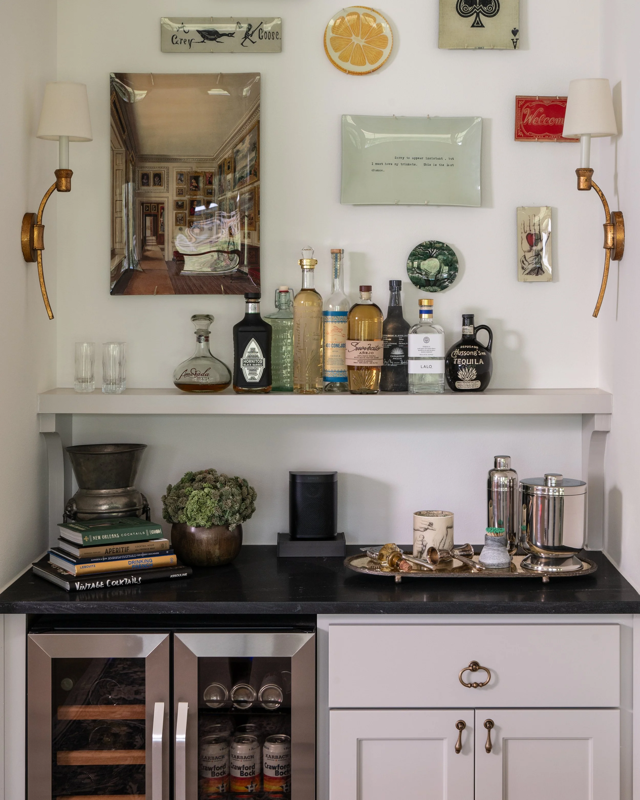 A bar area with liquor bottles on a white shelf, a black countertop with cocktail books, a potted plant, and cocktail accessories. Framed artwork, mirrors, and wall-mounted lamps decorate the white wall above.