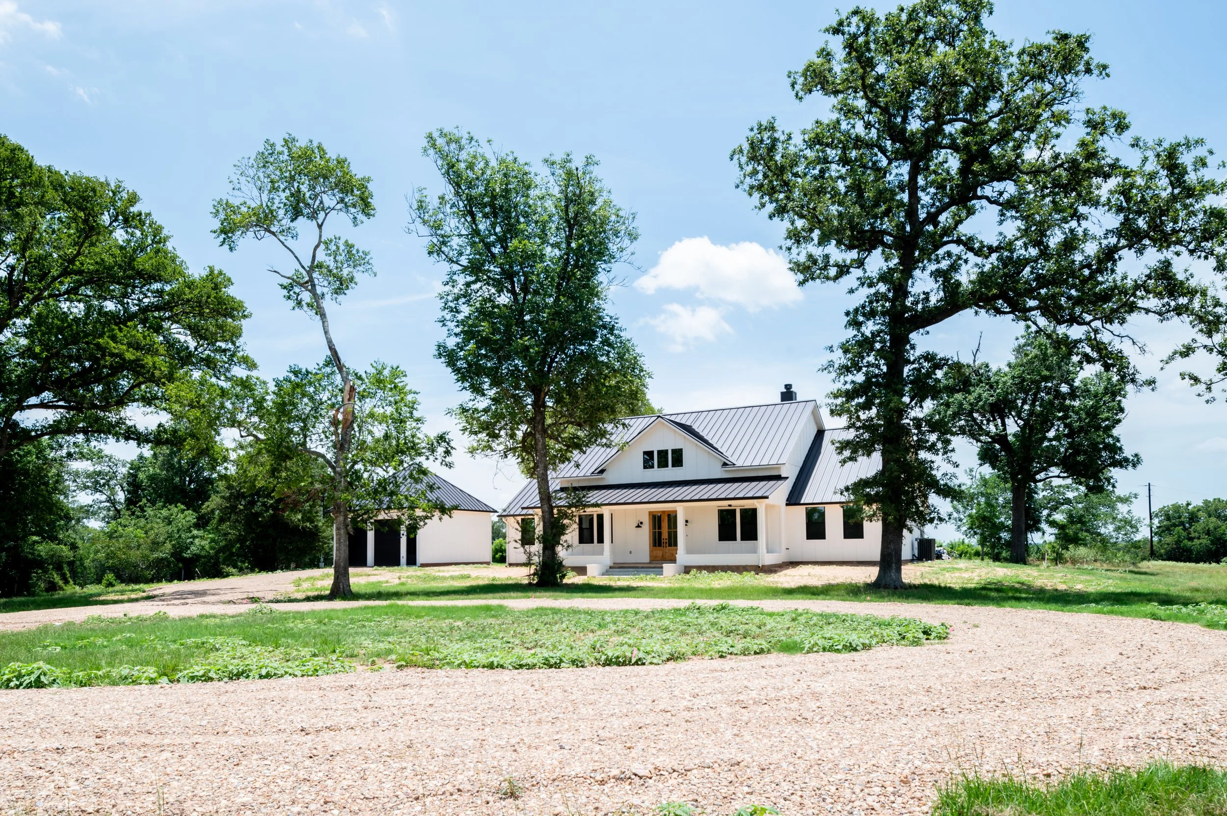 White house with a metal roof in a rural setting, surrounded by green trees and a gravel driveway, under a partly cloudy sky.