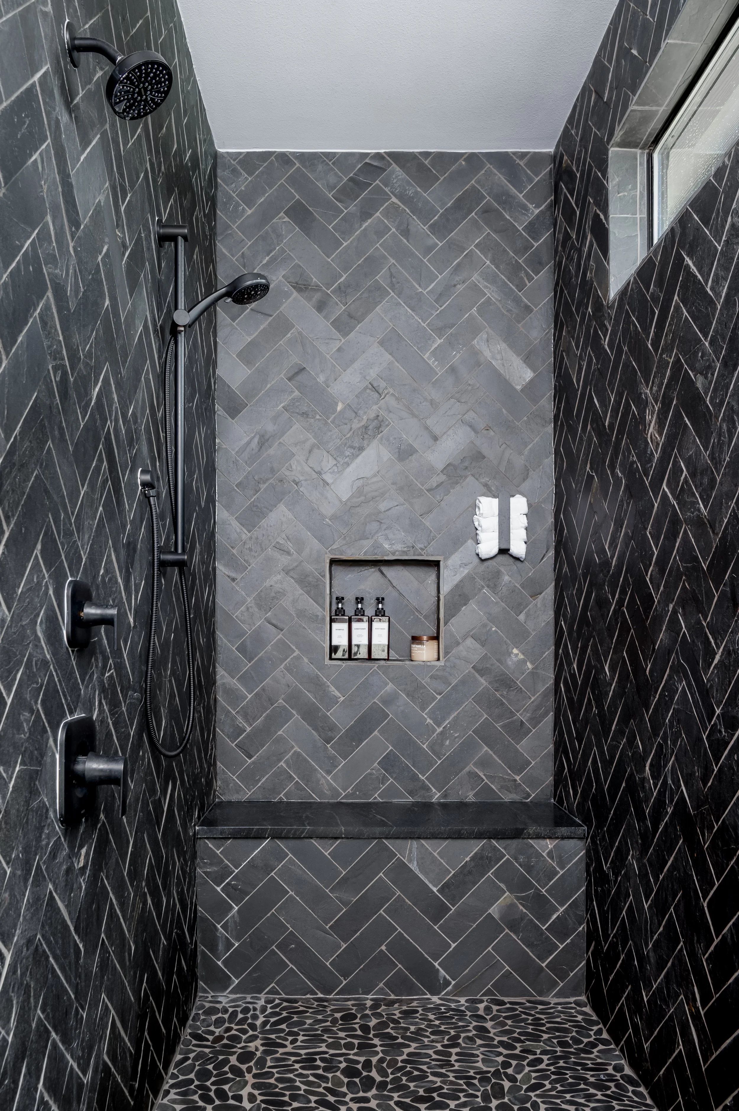 Modern shower with dark gray herringbone tiles on the walls, a pebble stone floor, and a built-in niche with toiletries. Two adjustable shower heads are mounted on the left wall, and a small window is near the top right corner.