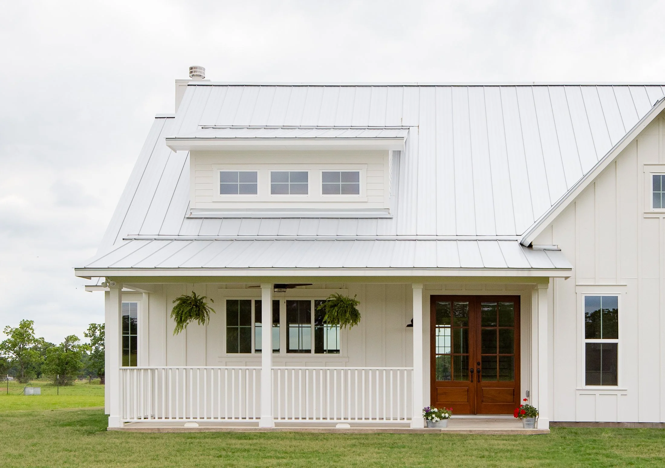White farm-style house with metal roof, wooden front door, front porch with hanging plants, and potted flowers at entrance. Green lawn and trees in background.