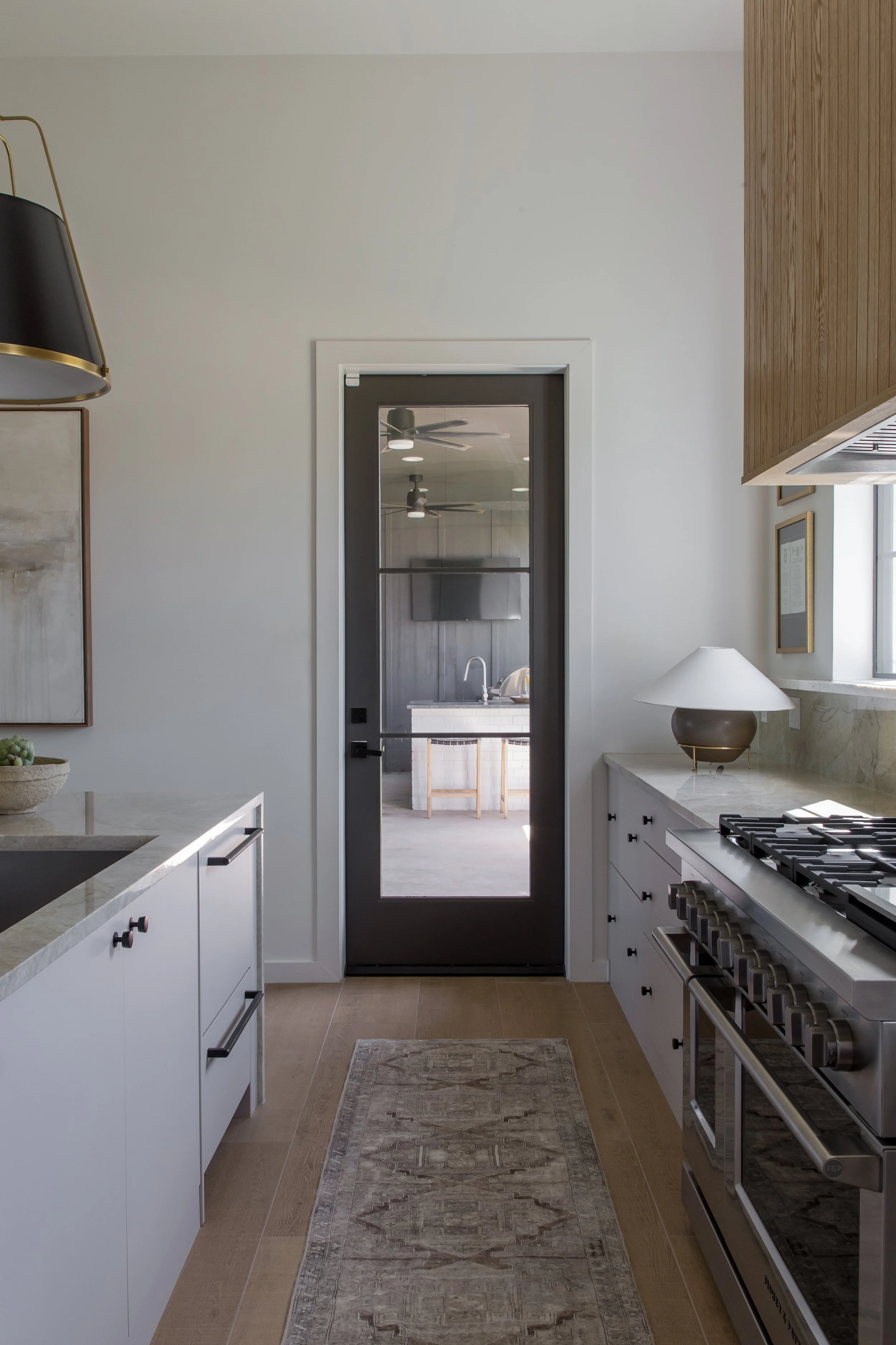 Interior view of a modern kitchen with white cabinets, a marble countertop, a black door with glass panel leading to another room, a gas stove, and a lamp on the counter. There is a rug on the wooden floor and a window with sunlight.