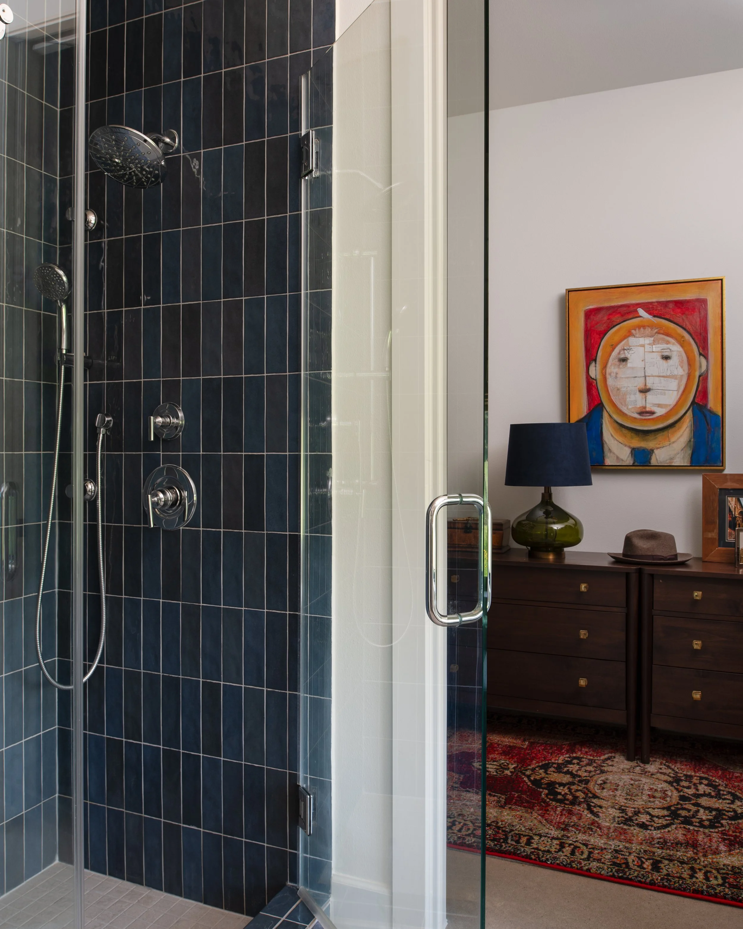 A bathroom shower with dark blue tiled walls and a glass door, adjacent to a living space with a wooden console, lamps, a hat, and a colorful artwork of a face wearing a lion mask.